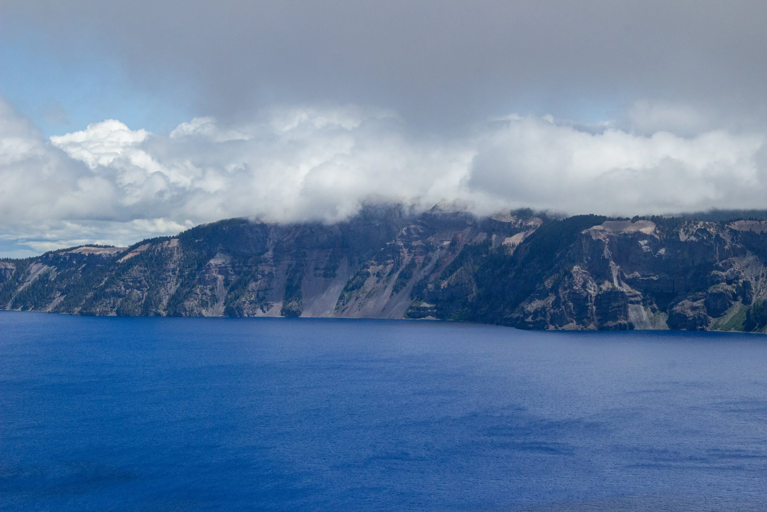 A large body of deep blue water in the foreground with rugged, forested mountain cliffs in the background beneath a cloudy sky.