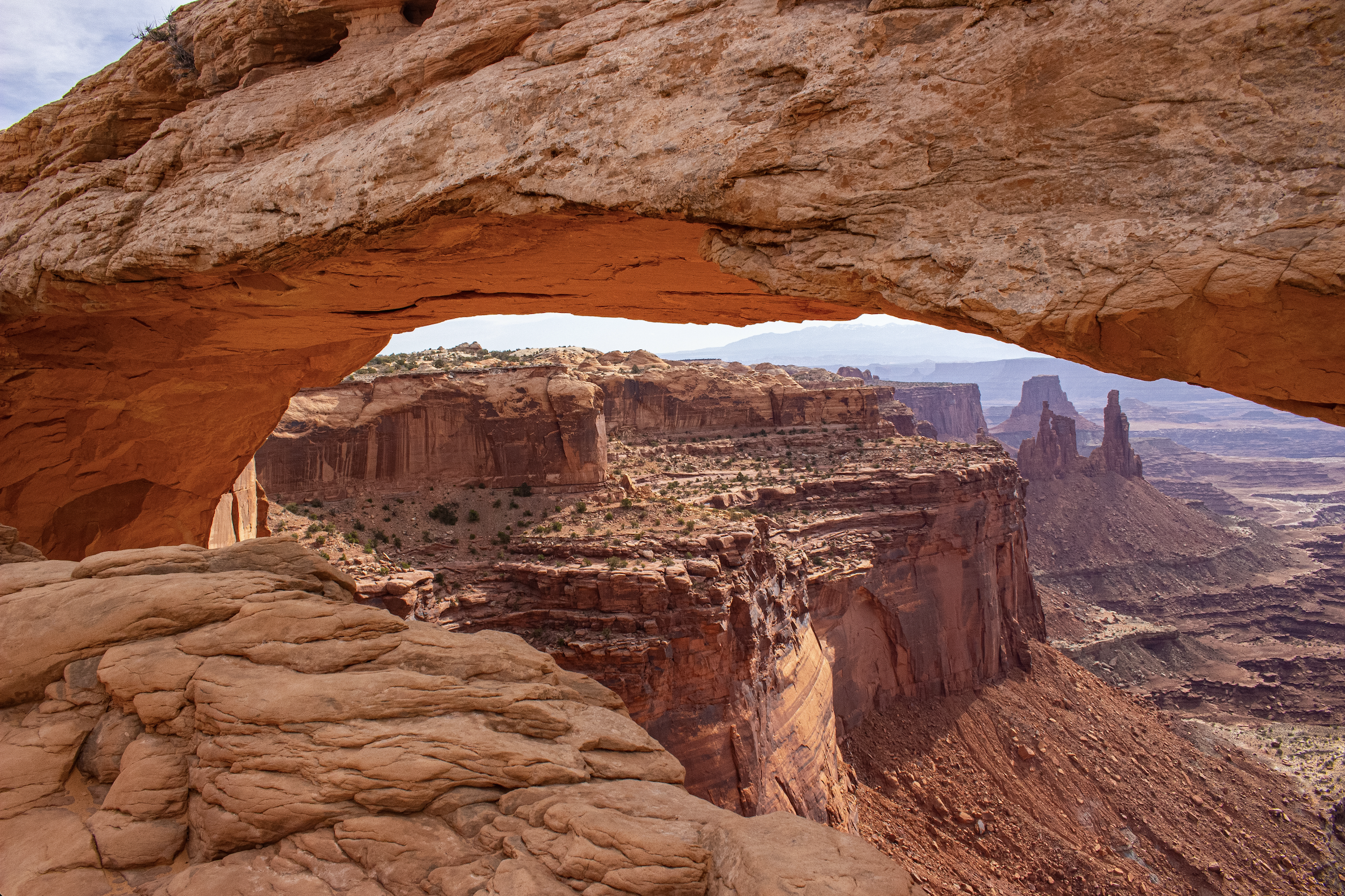 Rock formation with a natural arch overlooking a canyon landscape with cliffs and rock spires in the distance.