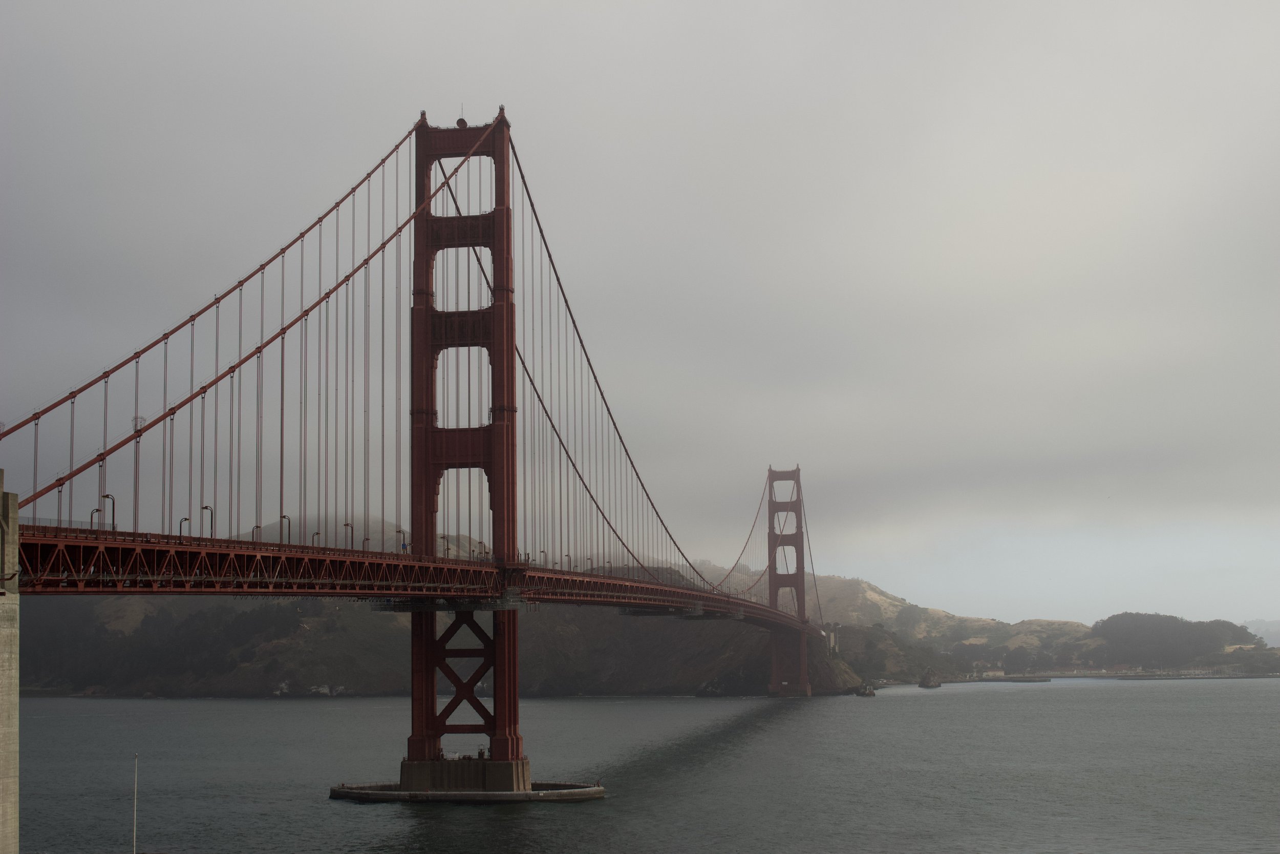 A view of the Golden Gate Bridge with its iconic red towers and cables, shrouded in fog over the water with hills in the background.