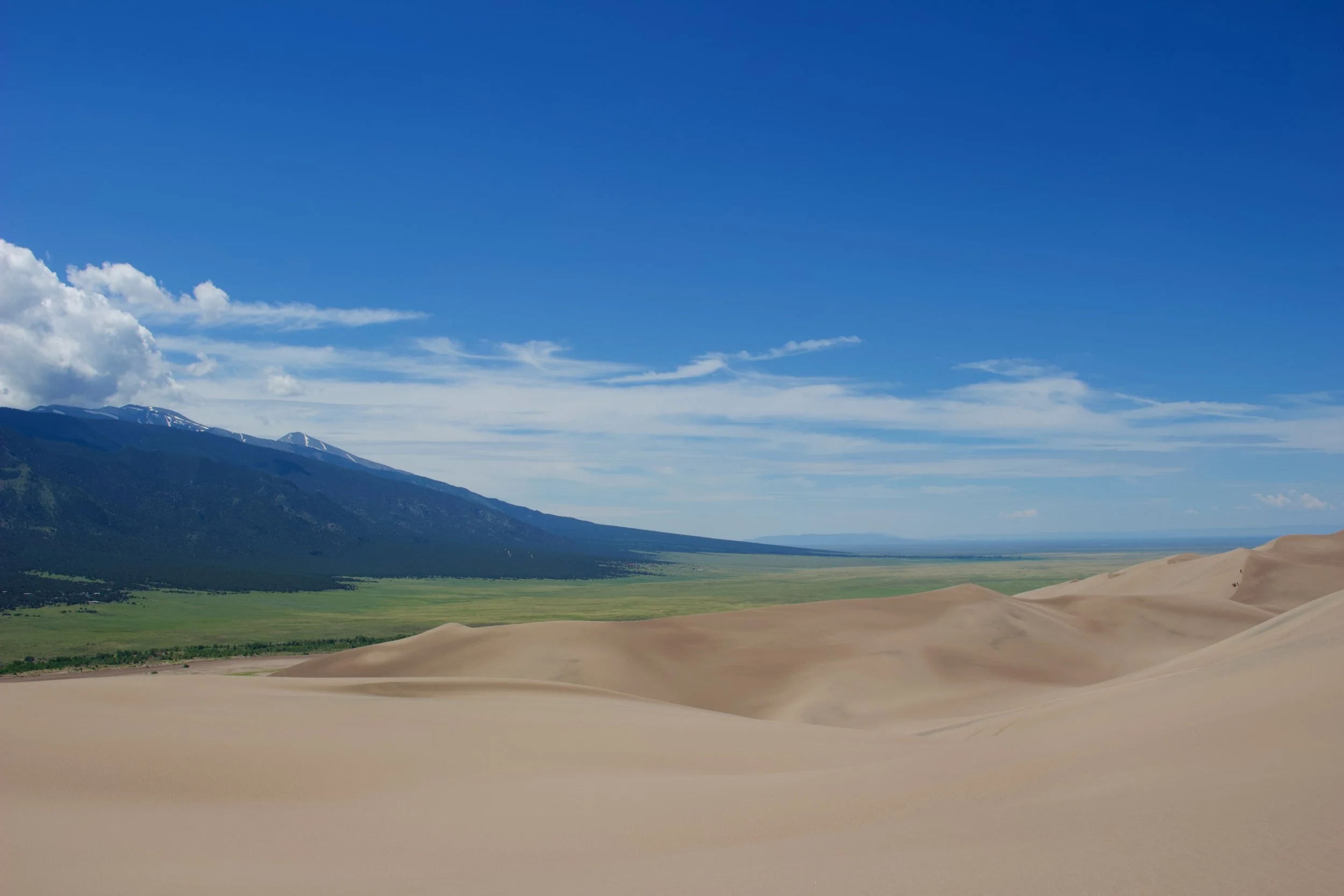 A landscape featuring sand dunes in the foreground, green plains and a mountain range with snow-capped peaks in the background, under a bright blue sky with some clouds.