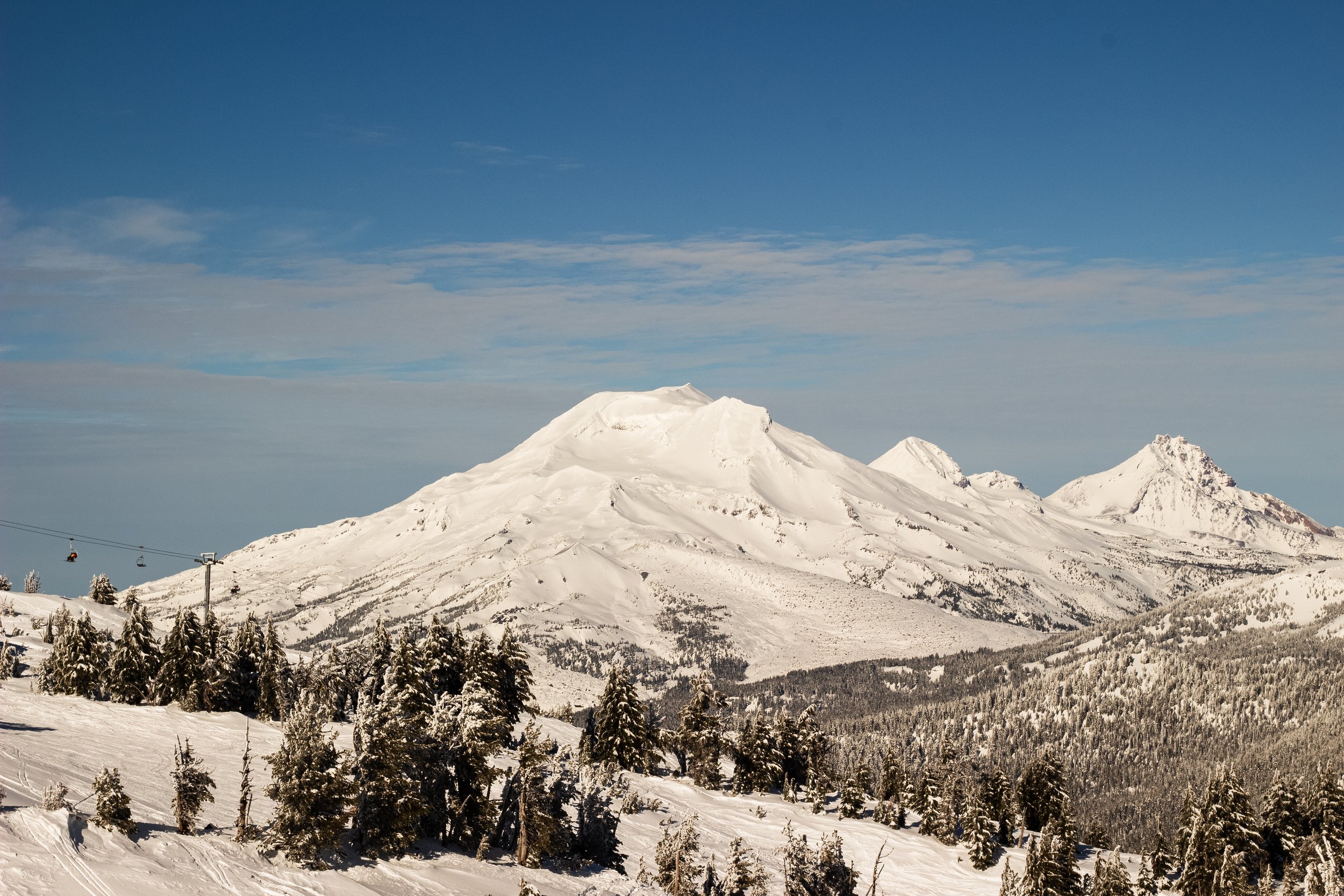 Snow-covered mountain range with a ski lift in the foreground and a blue sky with some clouds.