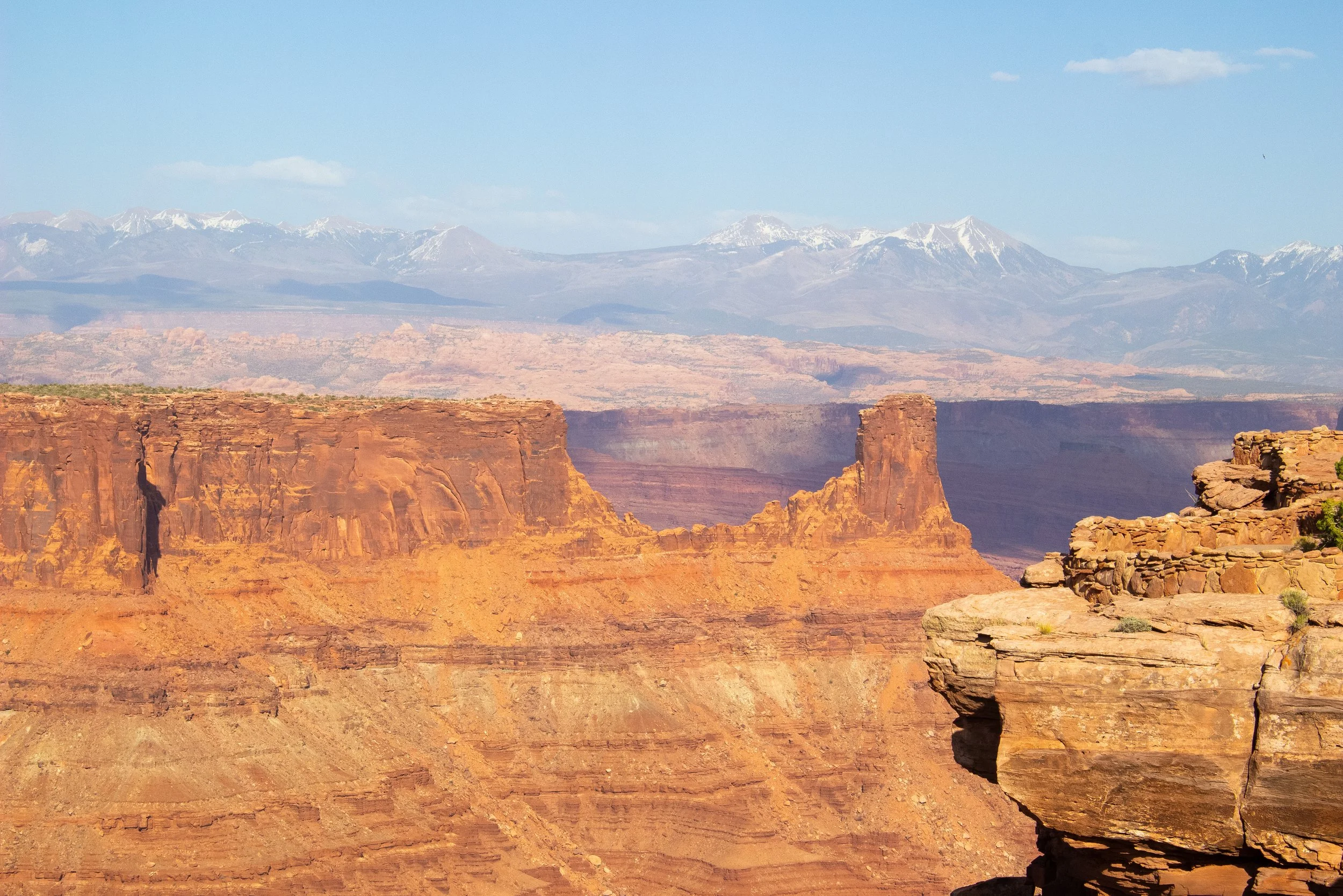 Scenic view of the Grand Canyon with reddish rock formations in the foreground and snow-capped mountains in the background under a blue sky.
