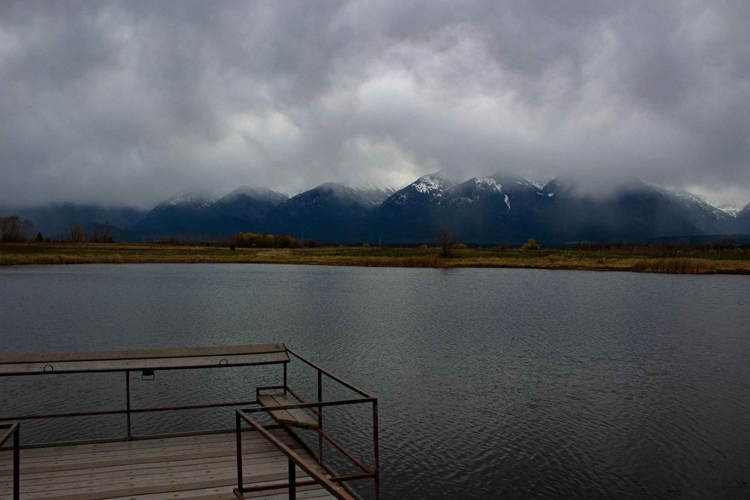 A wooden dock with metal railings extends into a body of water, with a mountain range partly shrouded in clouds in the background on a cloudy day.