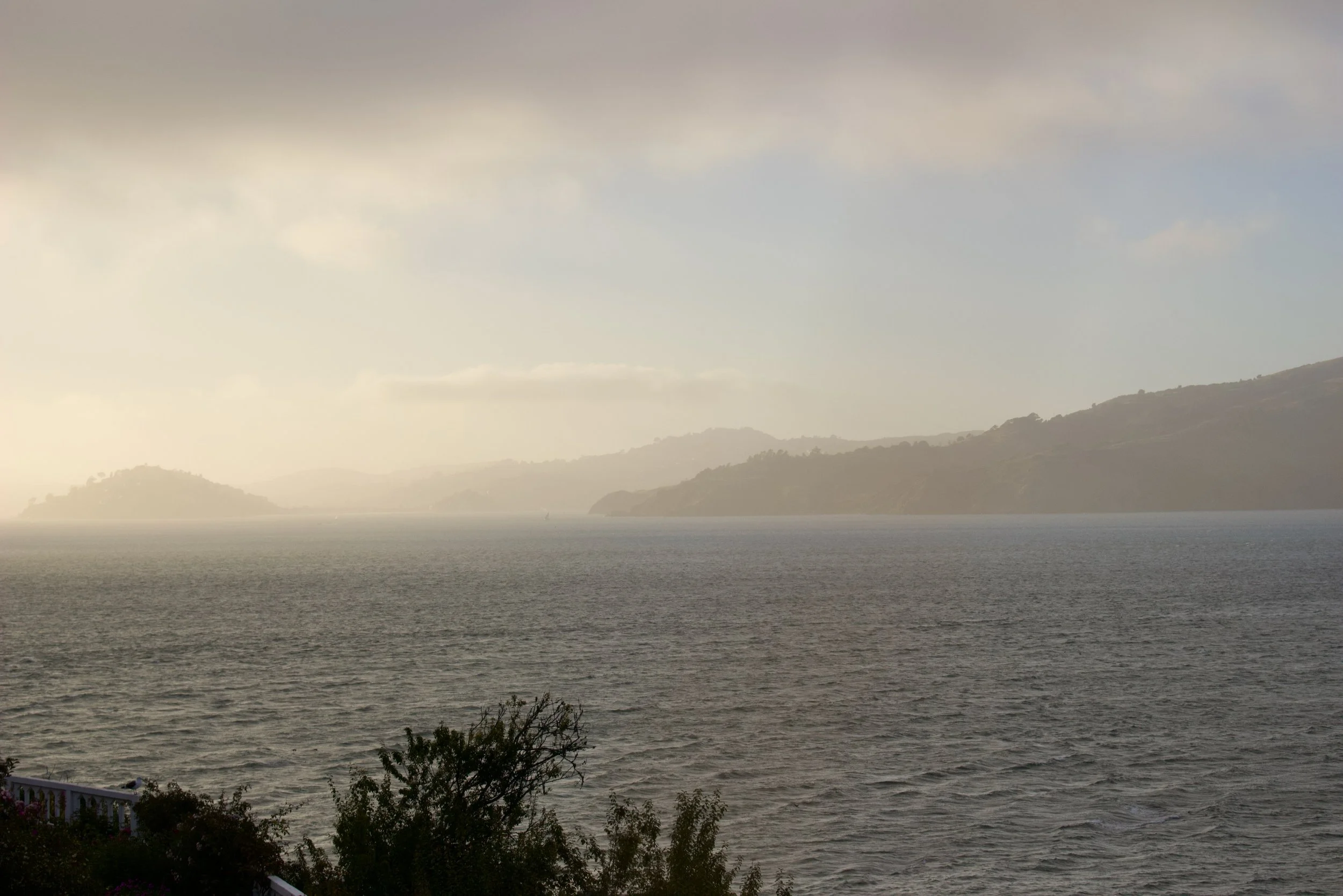 A view of a large body of water, possibly a lake or ocean, with distant hills or mountains in the background and a cloudy sky above.