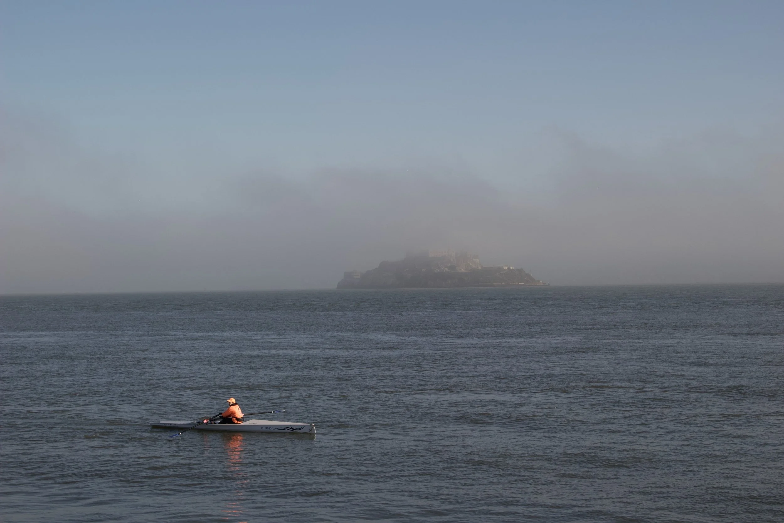 Person rowing a small boat on calm water with an island in the background under a hazy sky.