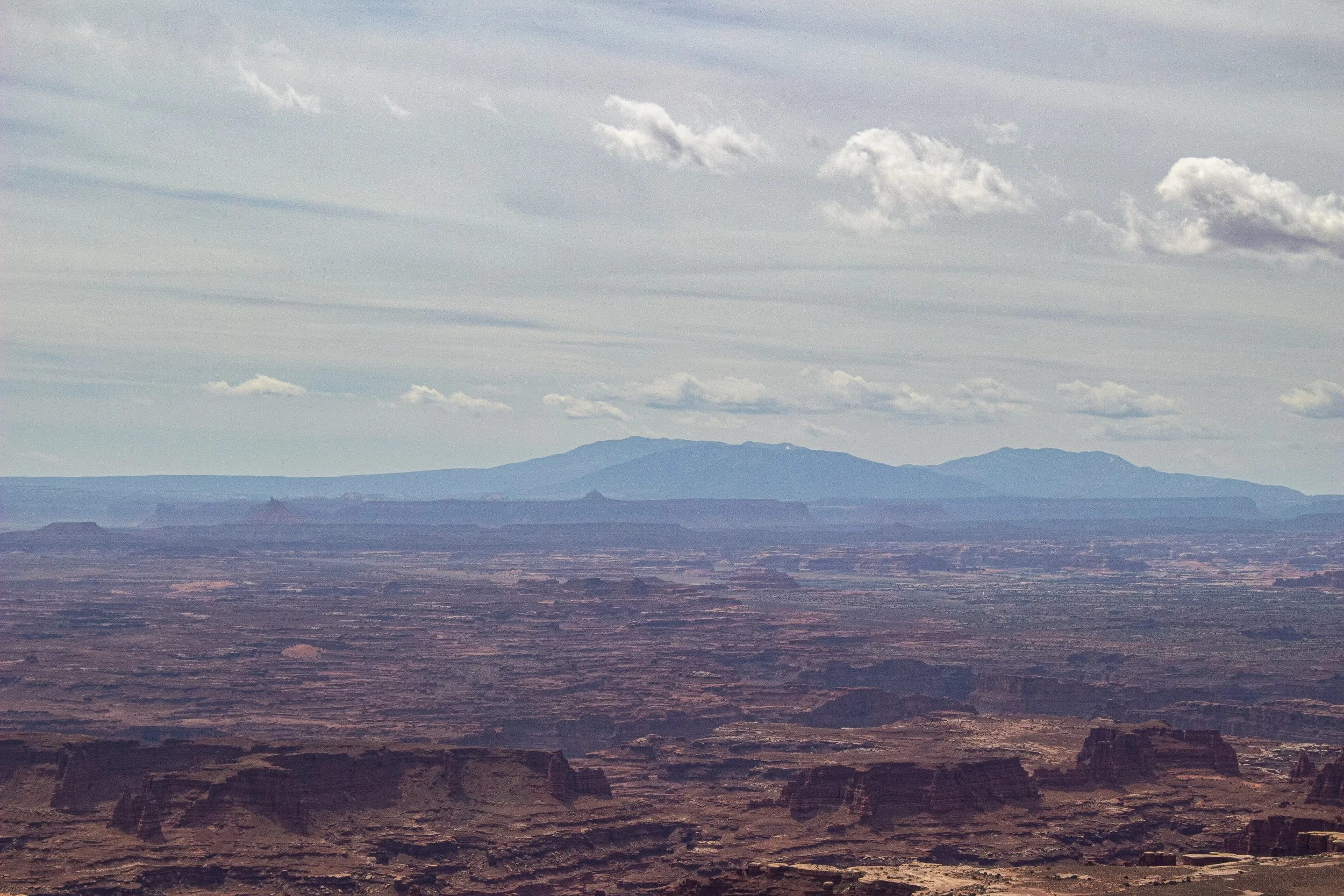 A landscape view of a vast desert with rugged terrain, mesas, and distant mountains under a cloudy sky.