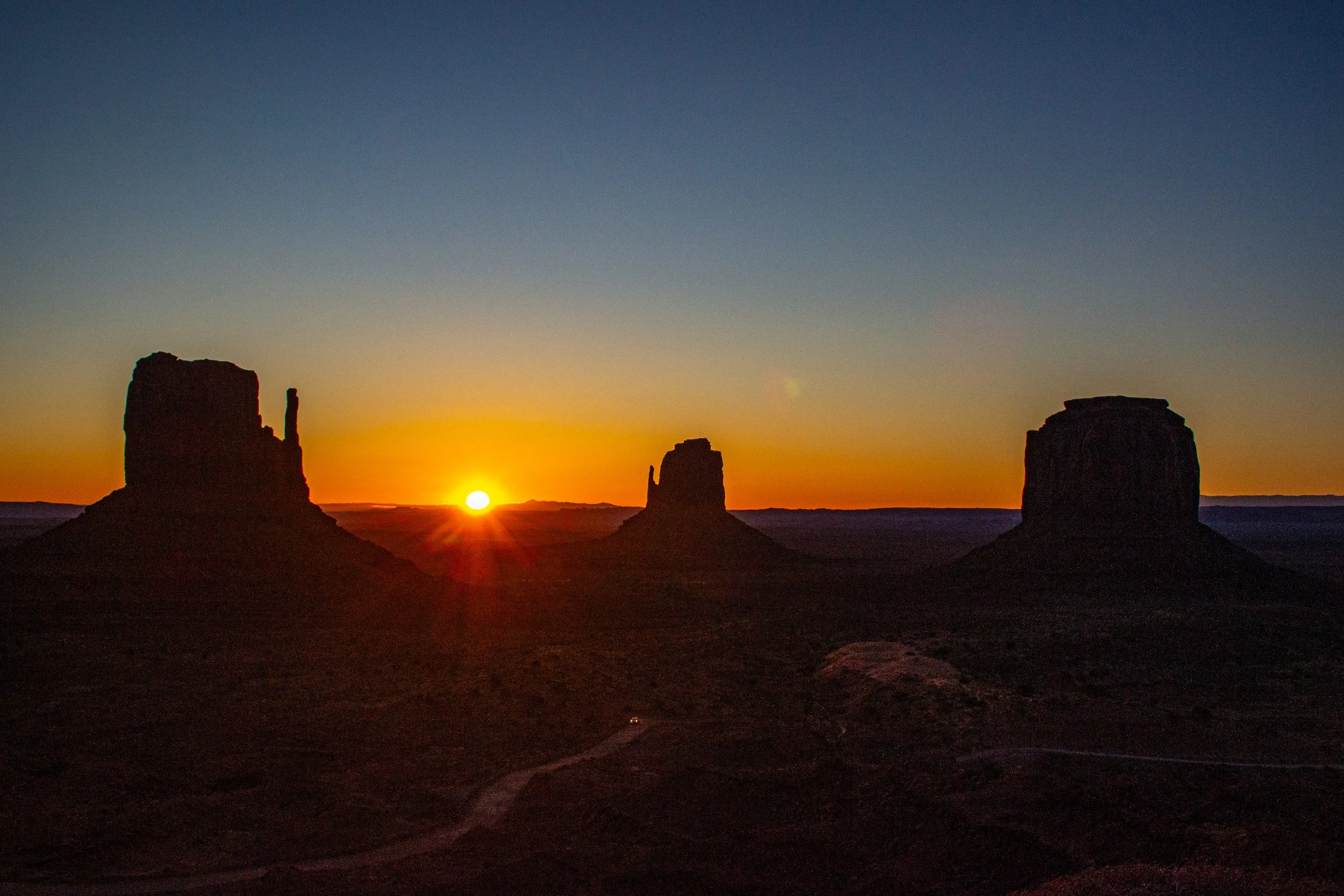 A desert landscape at sunset with three large rock formations silhouetted against a colorful sky, with the sun near the horizon.