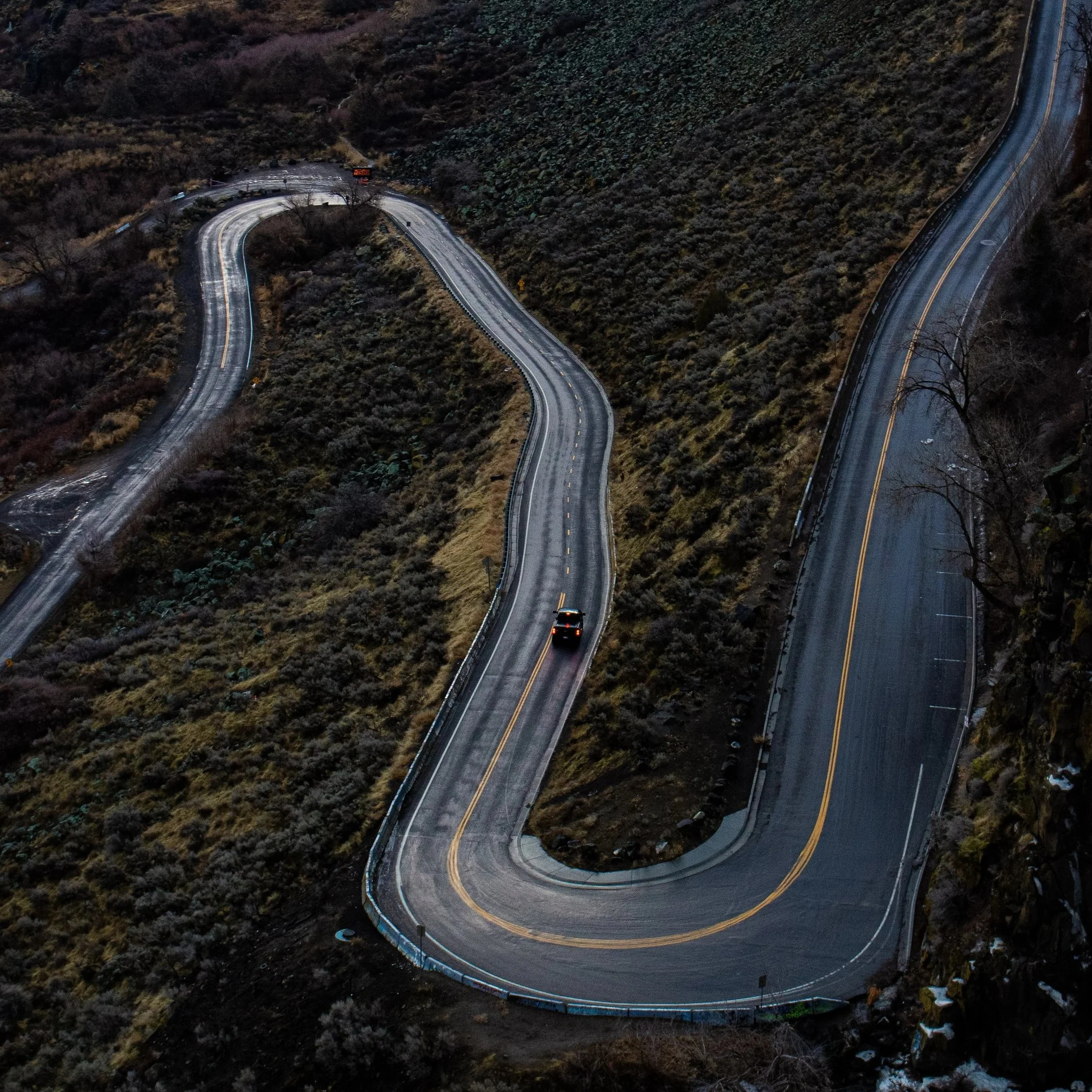 A winding mountain road at dusk with snow on the ground and cars traveling on it.