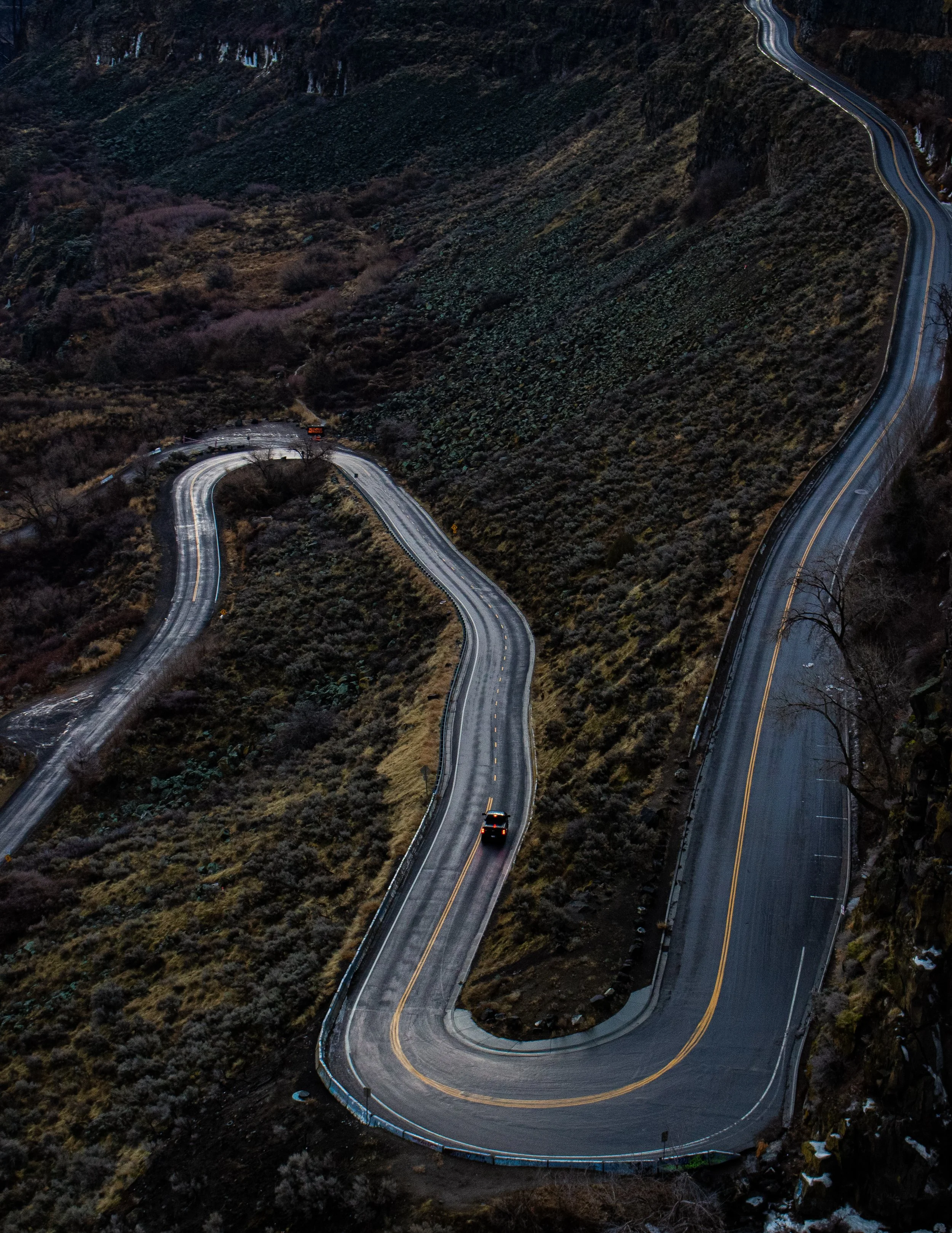 Twisting mountain road at dusk with two cars driving through the curves, surrounded by rugged terrain and sparse vegetation.