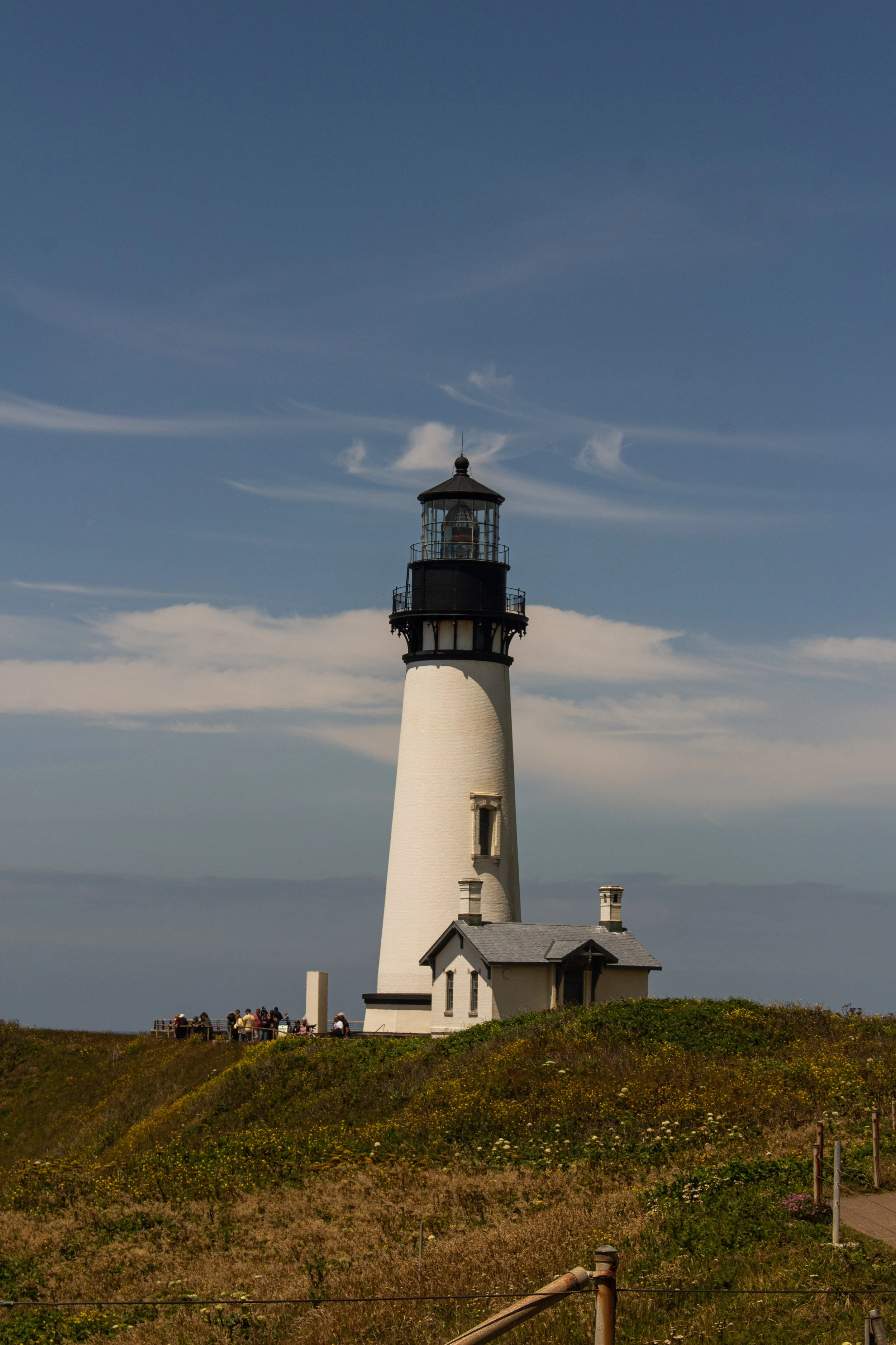 Lighthouse with white tower and black lantern room on a grassy hill, cloudy sky, and people walking near the lighthouse.