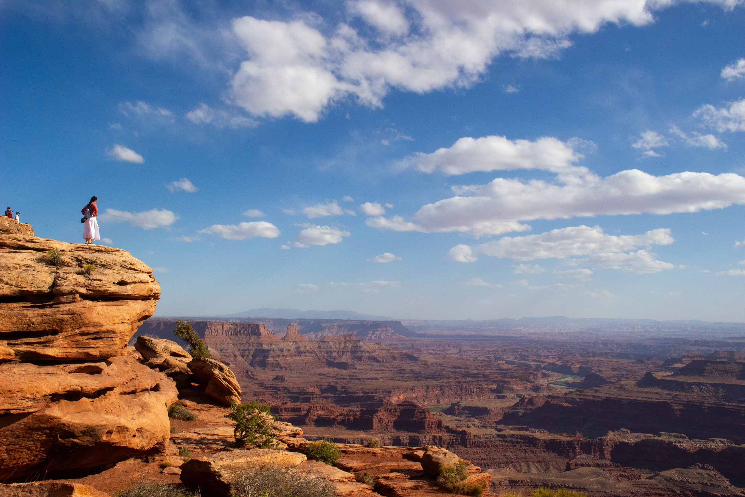 A person standing on the edge of a rocky cliff overlooking the Grand Canyon on a clear day with a blue sky and scattered clouds.