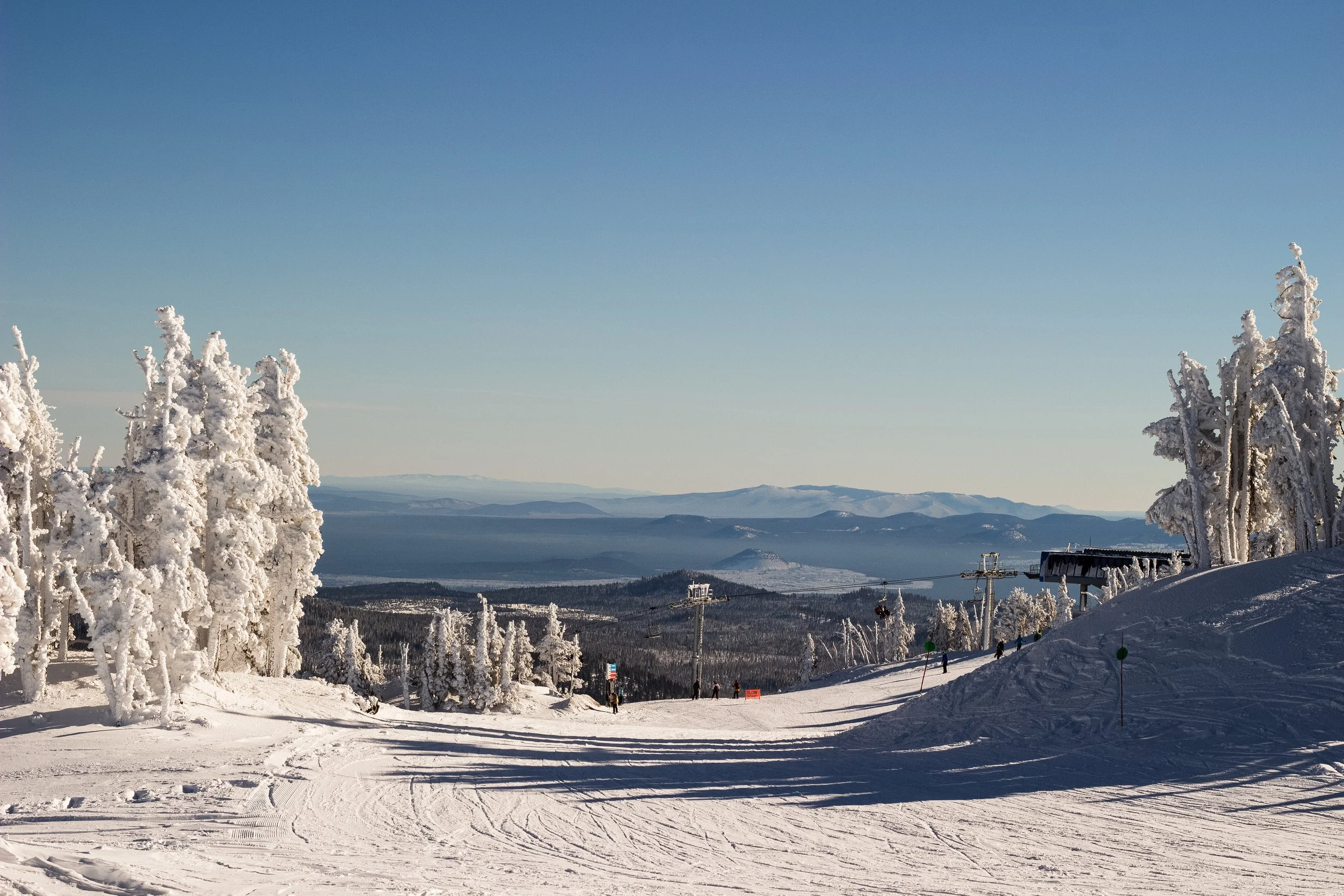 A snowy ski resort with snow-covered trees, ski lifts, and a mountain landscape in the distance under a clear blue sky.