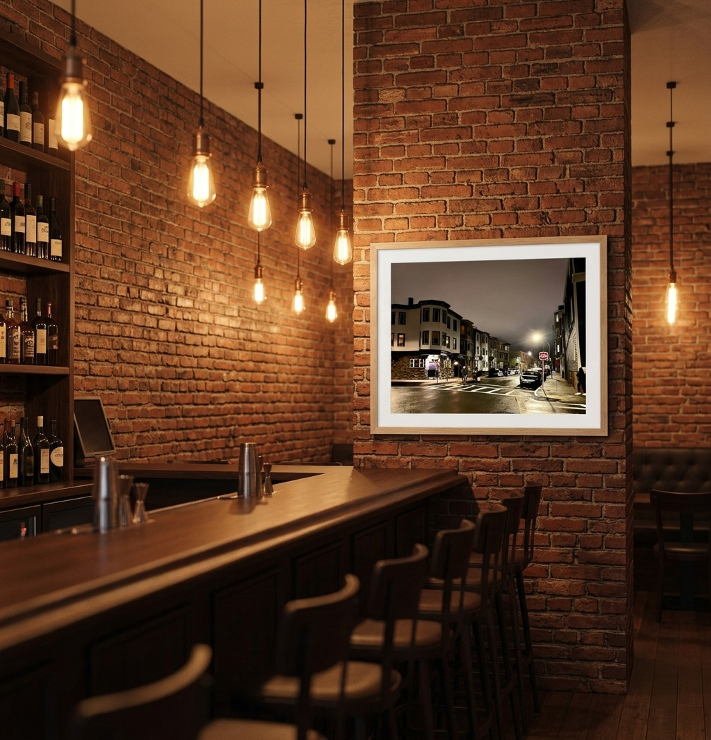 Interior of a cozy restaurant or bar with exposed brick walls, a row of hanging Edison bulbs, a wooden bar counter with barstools, shelves with bottles of wine, and a framed photograph of a city street at night on the wall.