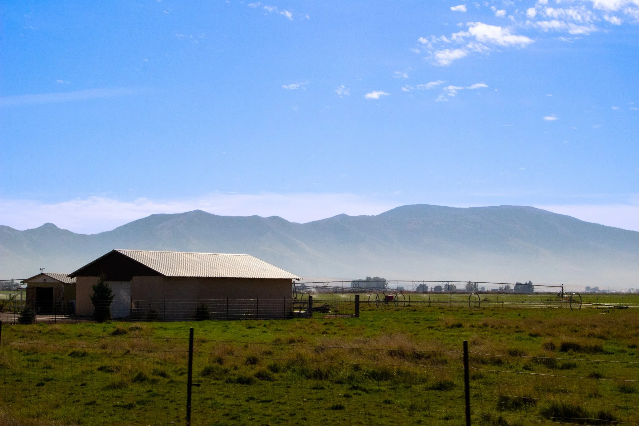 A rural landscape with a small building with a metal roof, a fenced green field, and distant mountains under a blue sky with scattered clouds.
