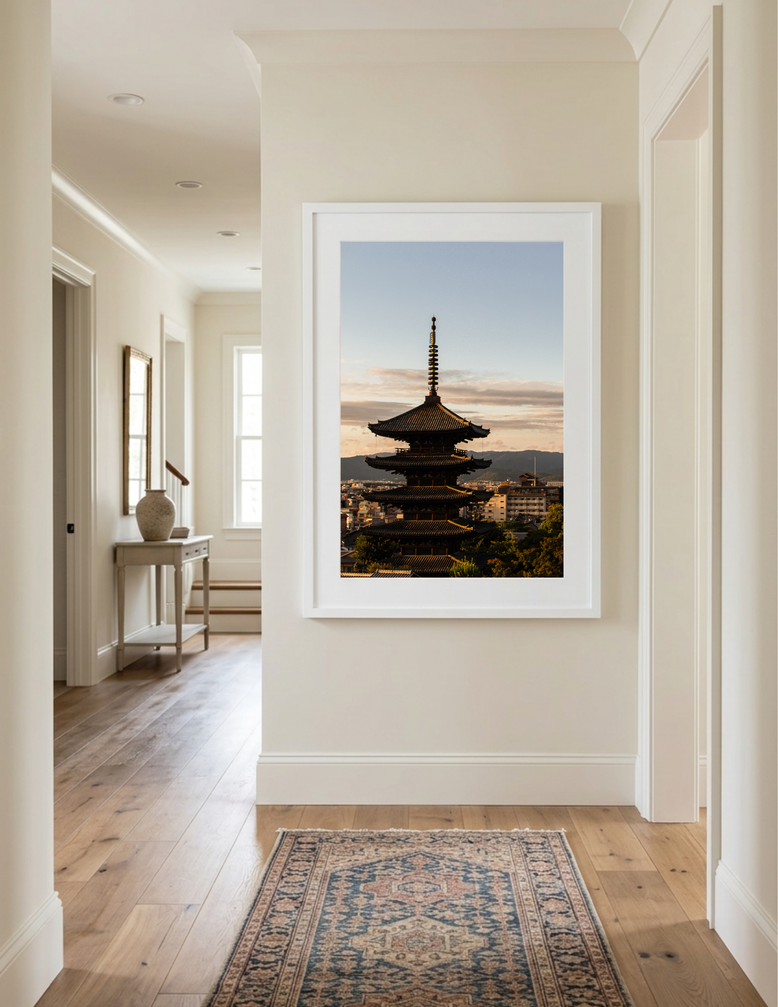 Interior view of a hallway with wooden flooring, a runner rug, a framed picture on the wall, a white console table with a decorative vase, and a large window in the background. There is a large framed photo of a pagoda-style building and cityscape outside the doorway.