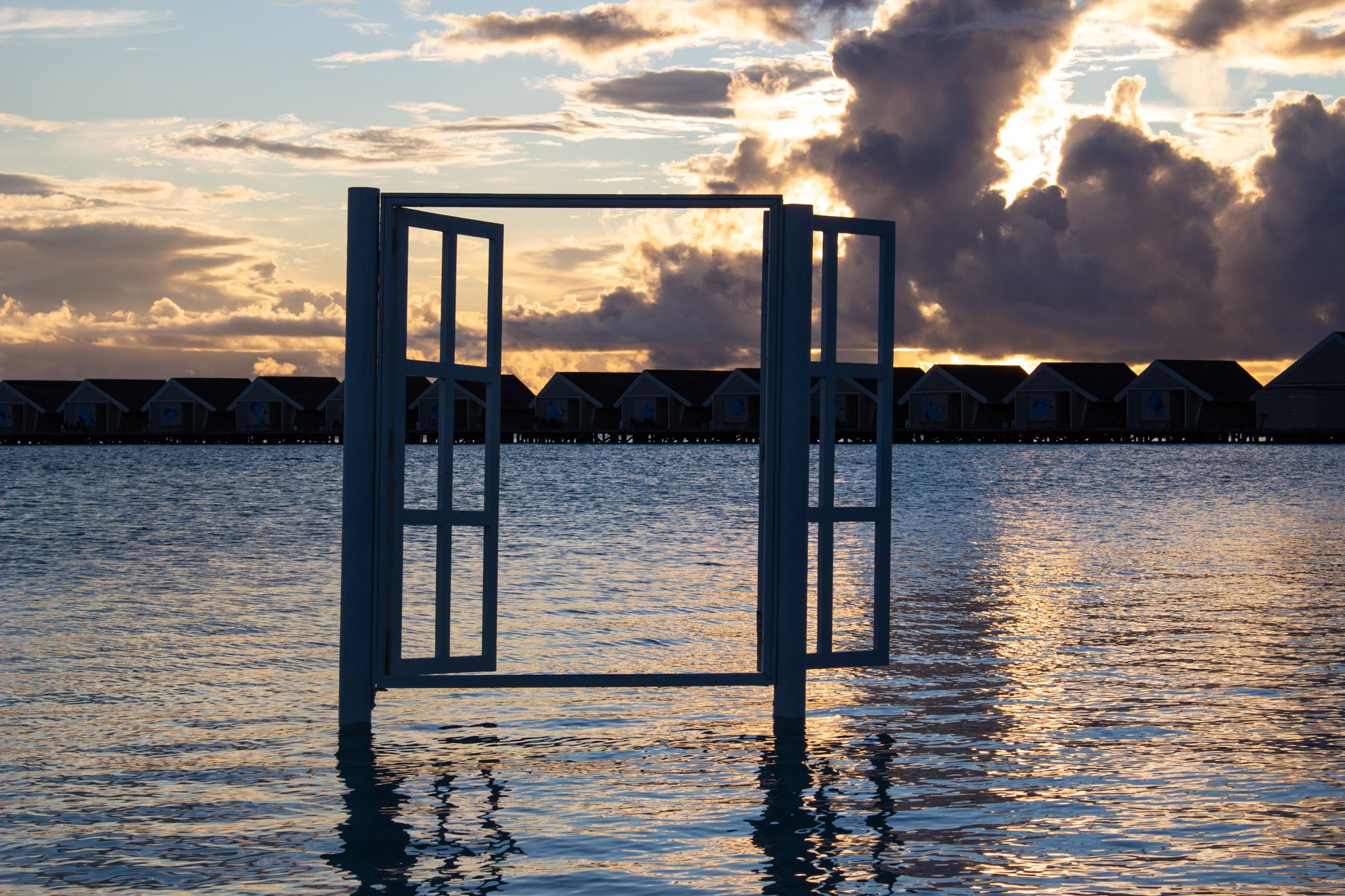 Open window frame overlooking water with houses on stilts and cloudy sky at sunset.