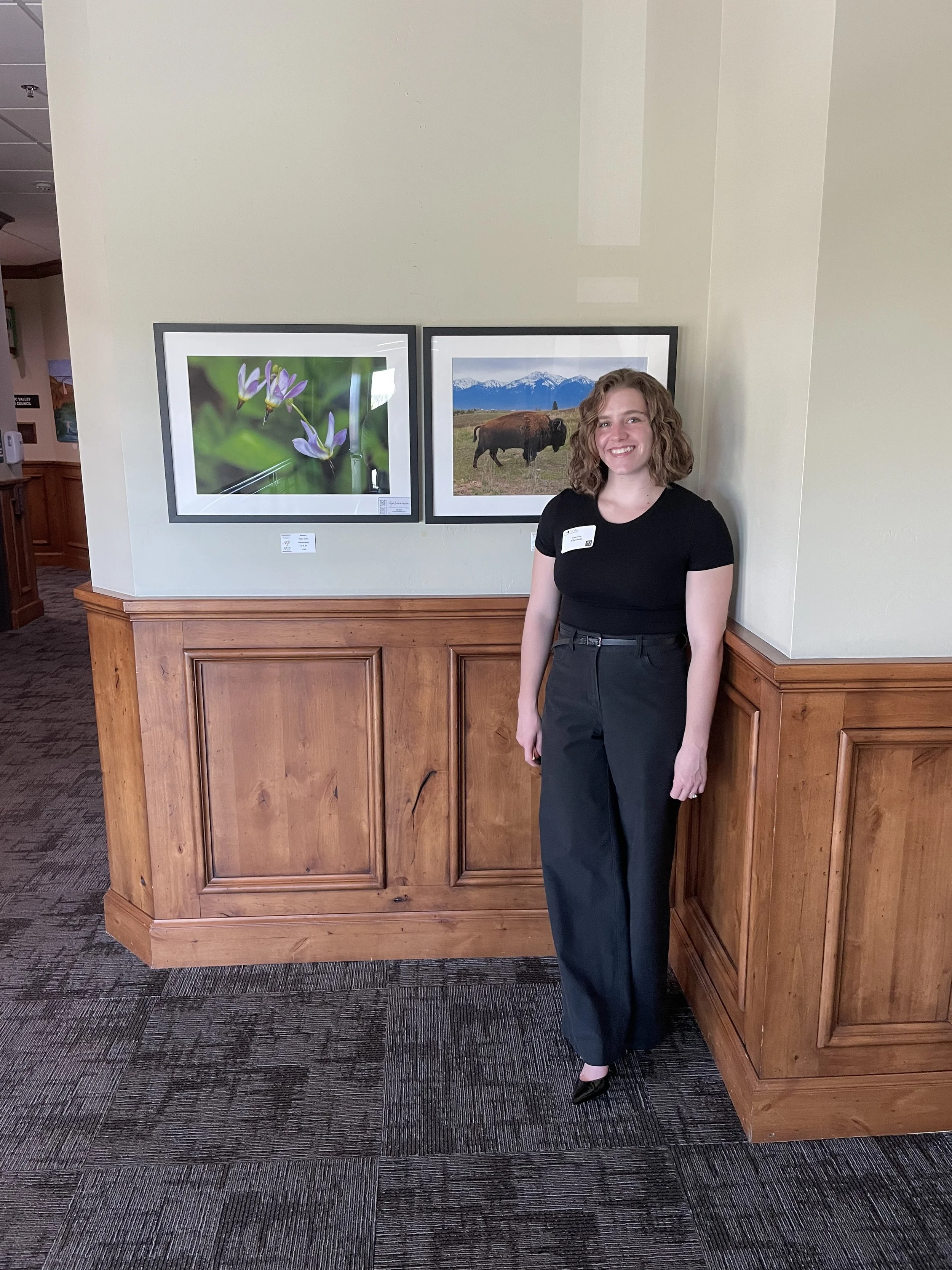 A young woman with wavy, shoulder-length brown hair smiling, wearing a black t-shirt, black high-waist pants, and black heels, standing next to a wooden reception counter in front of two framed landscape photographs on the wall.