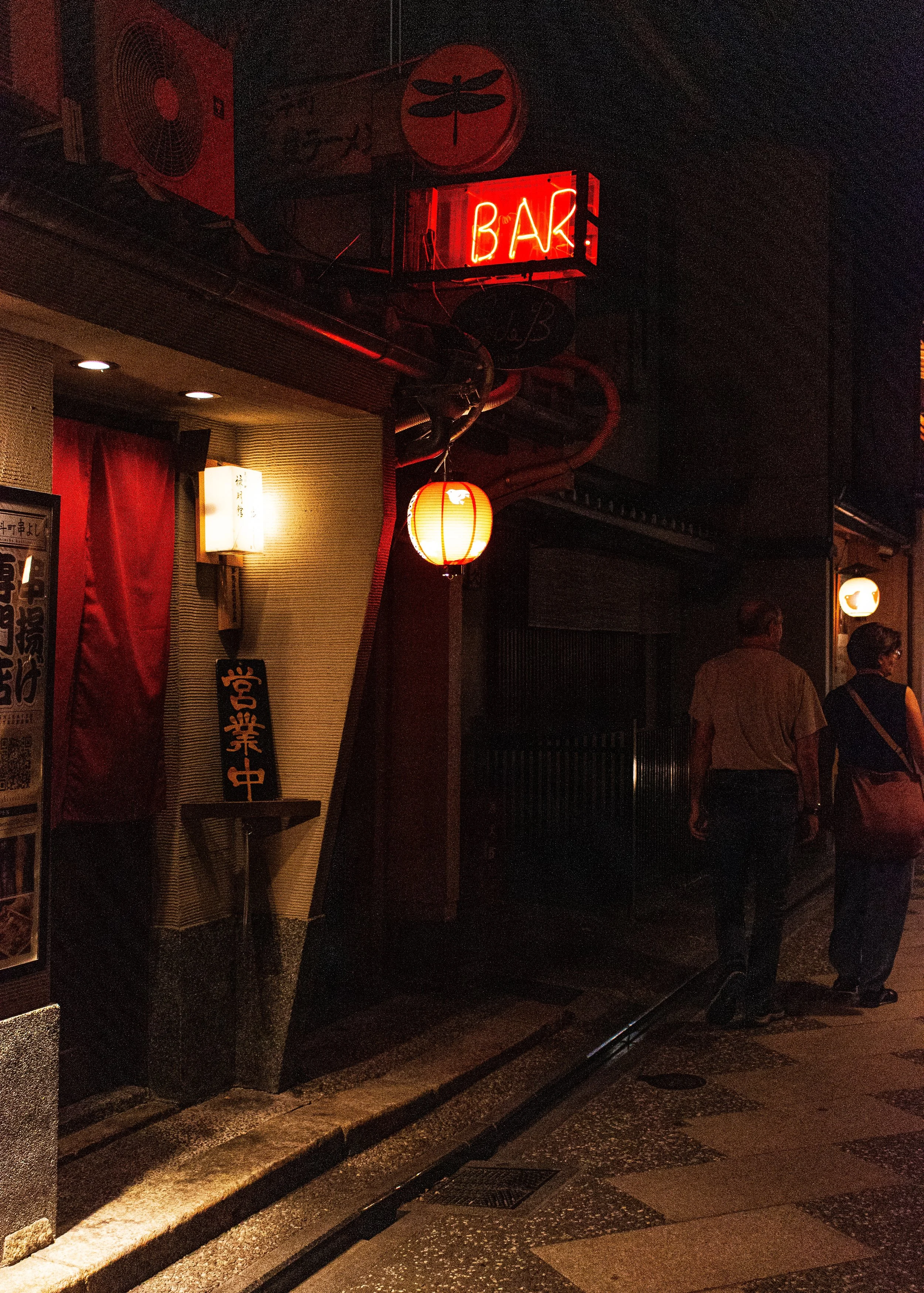 Night scene outside a Japanese restaurant with a red paper lantern and a neon sign that reads 'BAR.' The restaurant's entrance is partially covered by a curtain, and a couple of people are walking past on the sidewalk.