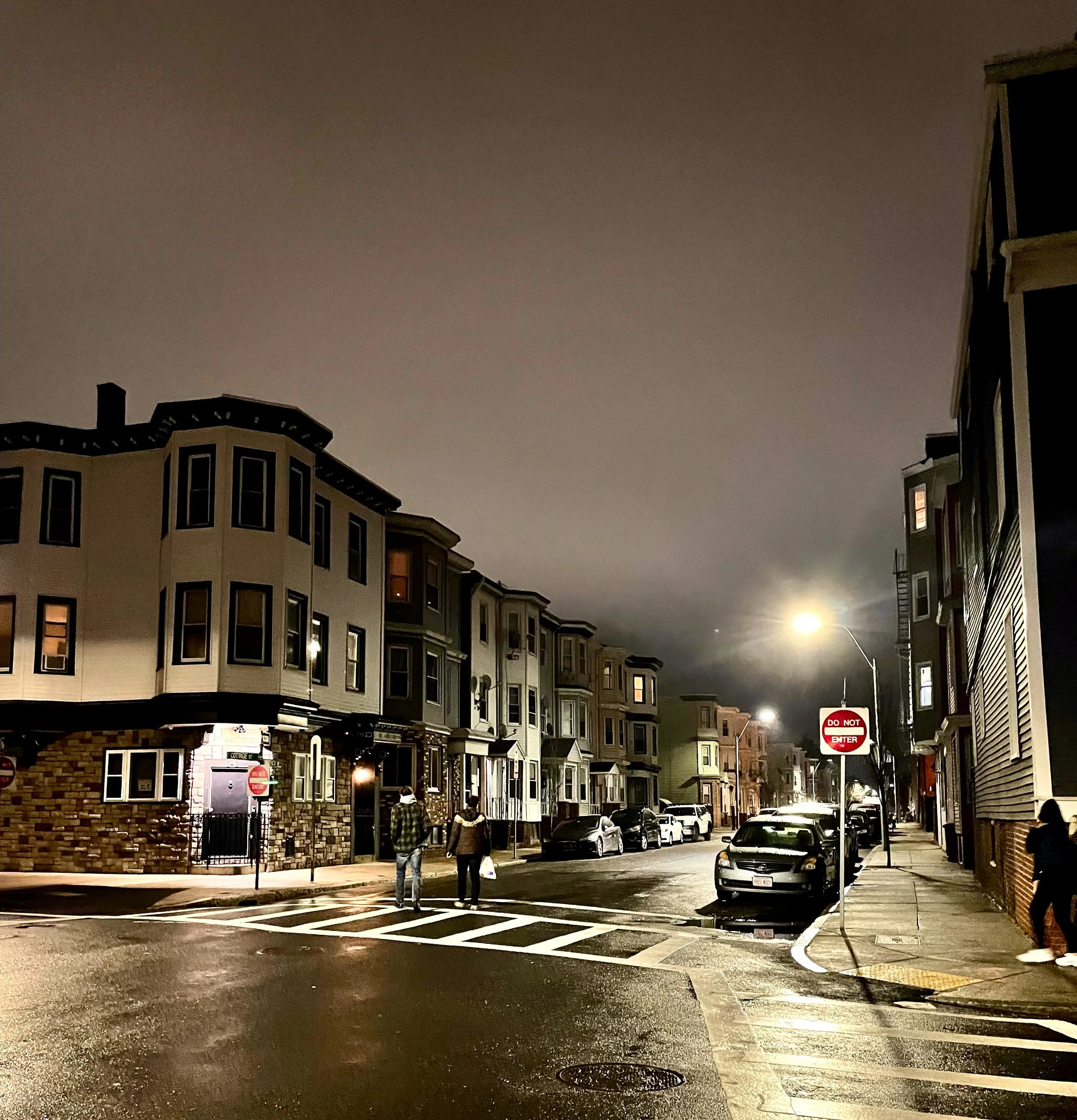 Night scene of a city street with buildings, cars parked along the street, and a streetlamp illuminating the area. Two people are walking on the sidewalk near a 'Do Not Enter' sign.