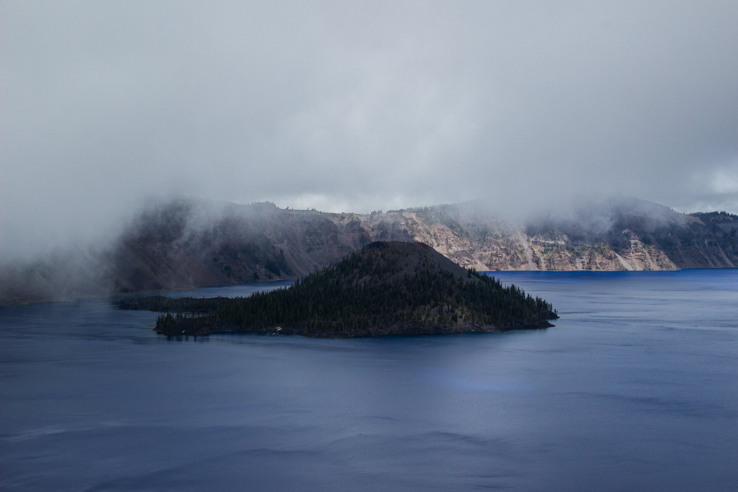 A misty mountain landscape with a large body of water in the foreground, forested hills, and fog-covered peaks.