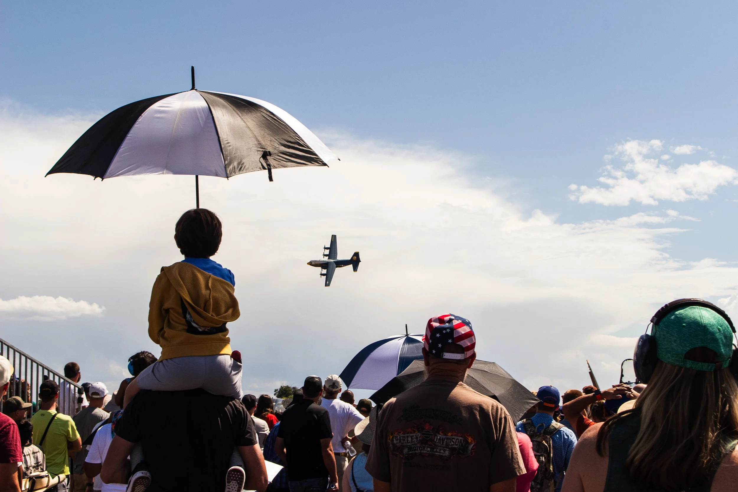 Crowd gathered outdoors under umbrellas, with a person riding a small airplane model in the sky.