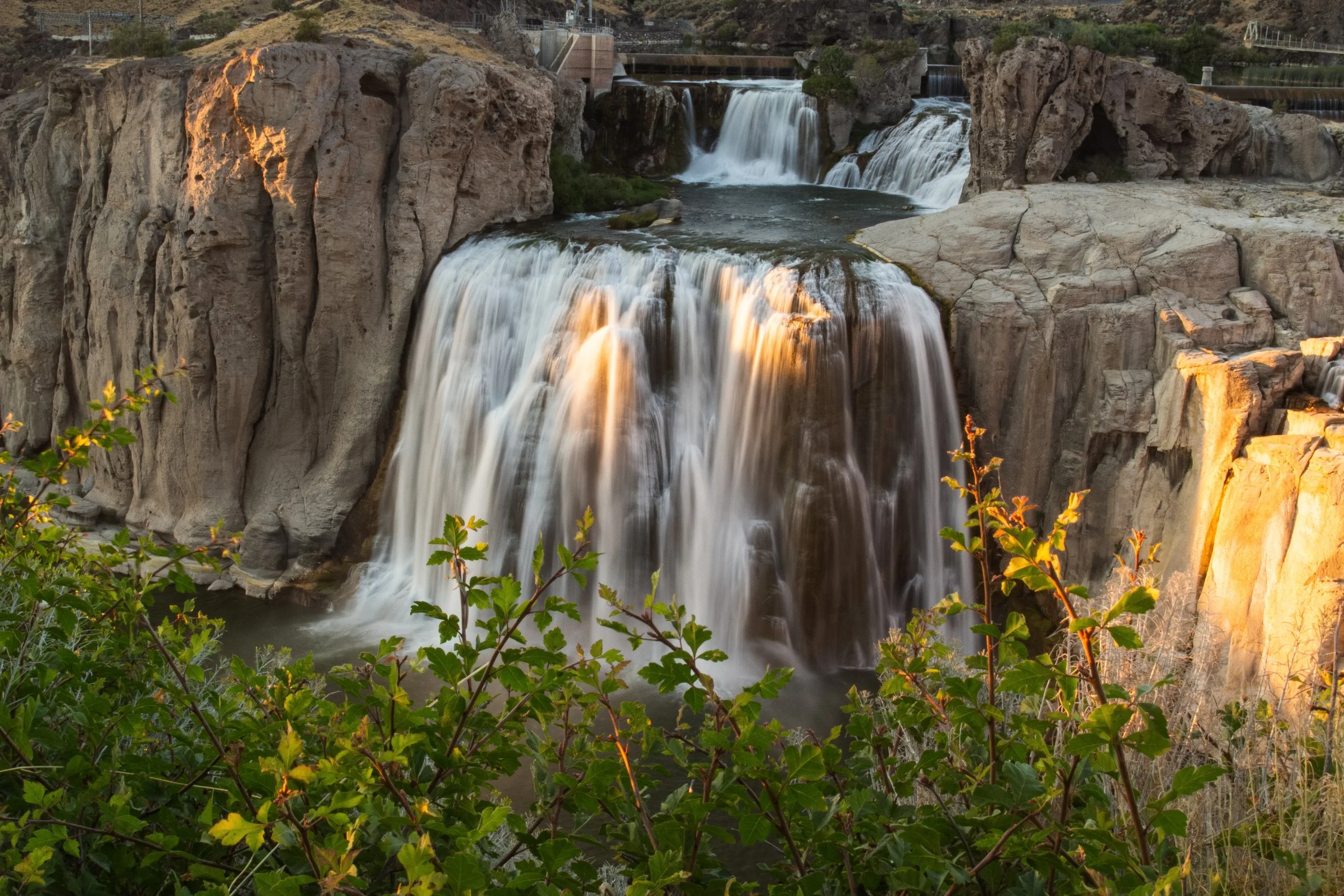Waterfall cascading over rocks in a canyon with greenery in the foreground and power plant structures in the background.