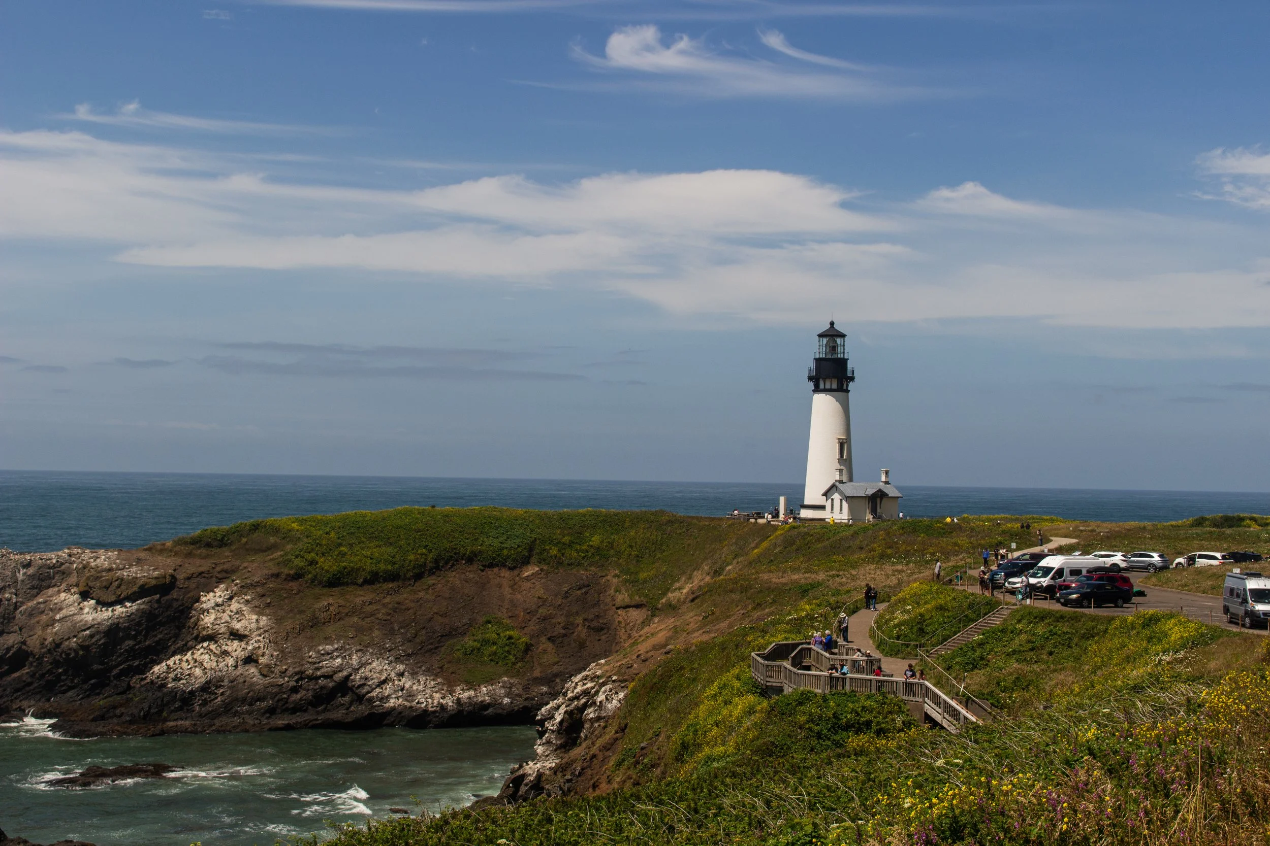 A scenic coastal scene with a white lighthouse near the ocean, surrounded by visitors walking along pathways and grassy areas, under a partly cloudy sky.