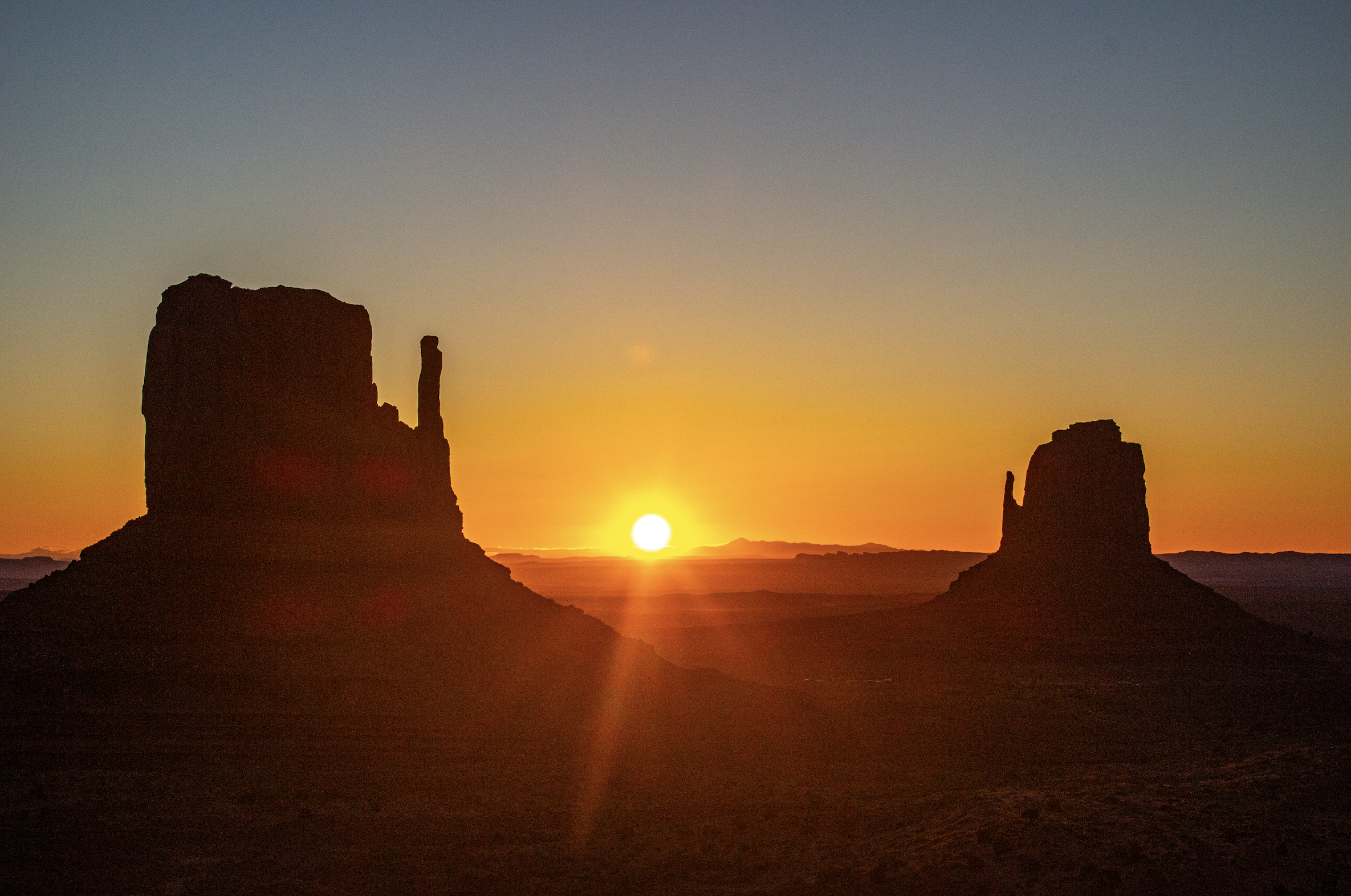 A desert landscape during sunset with large rock formations silhouetted against the colorful sky.