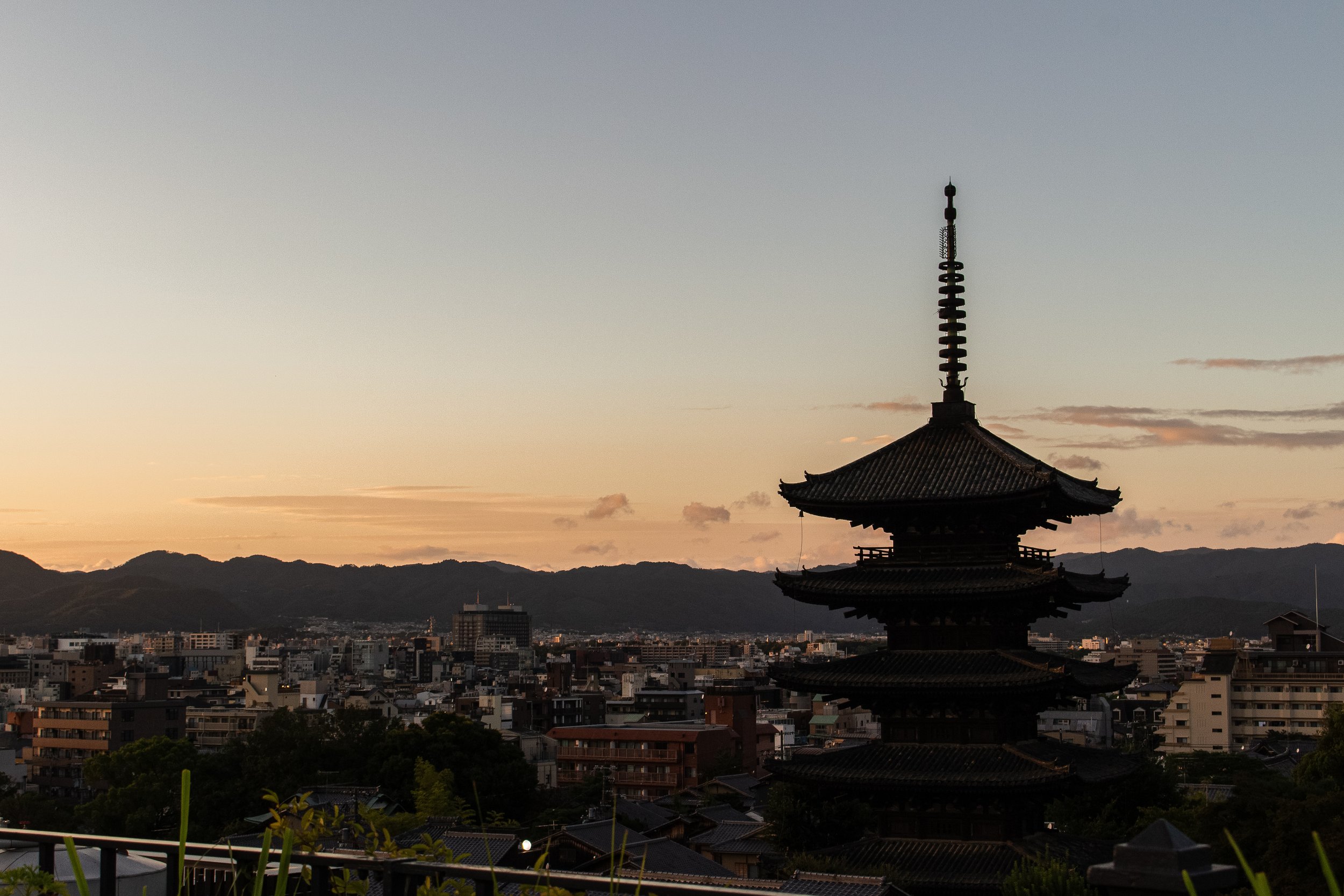 A traditional multi-tiered Japanese pagoda with a mountain range and a cityscape in the background during sunset.