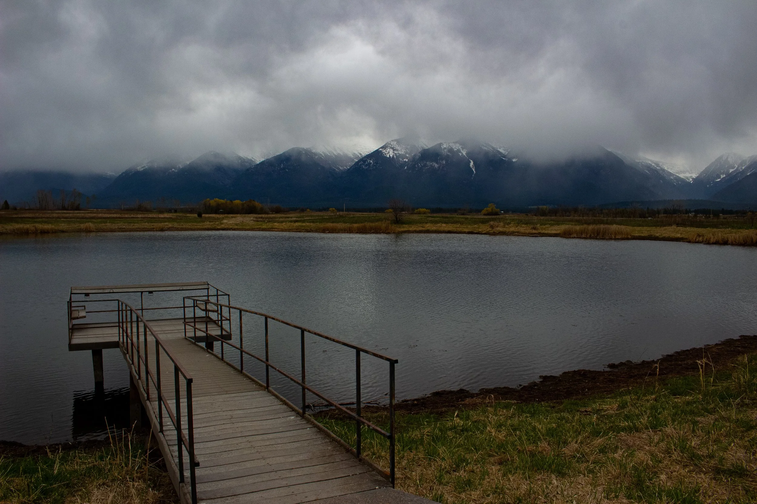 A cloudy day at a lake with a dock extending into the water, surrounded by grassy shores and mountains in the background.