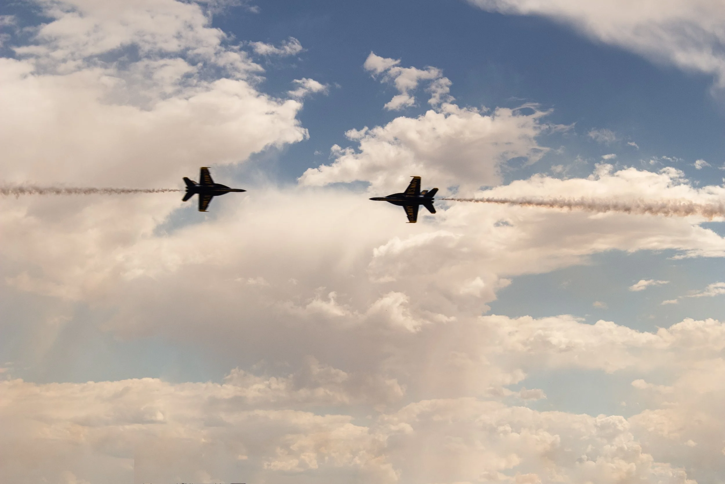 Two fighter jets flying in the sky, leaving smoke trails behind them, with clouds and blue sky in the background.