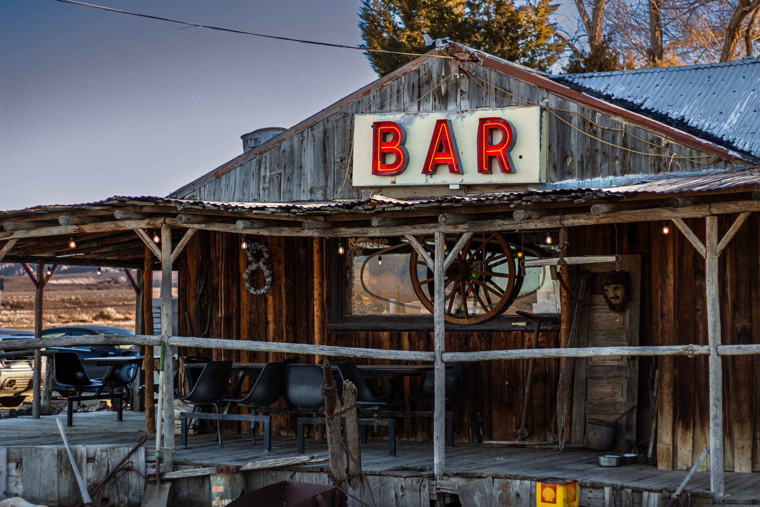 Rustic outdoor bar with a red neon sign that says 'BAR' on a wooden building with a metal roof, surrounded by trees and string lights.