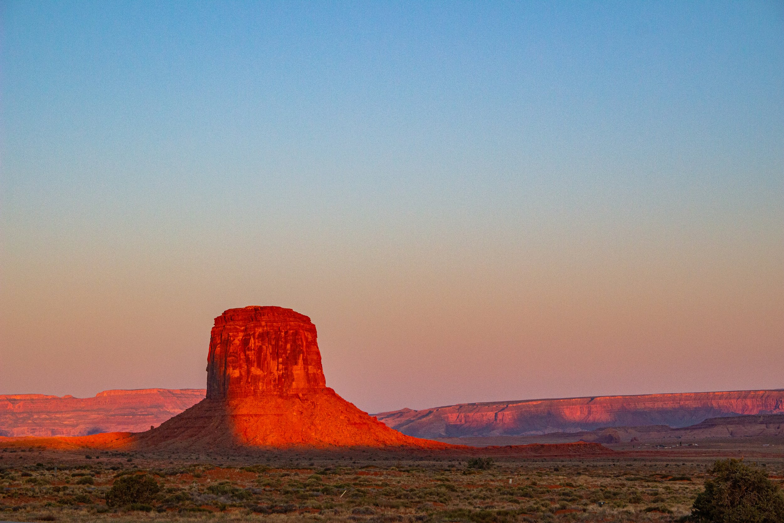 A large flat-topped red rock formation in a desert landscape during sunset with a clear sky.