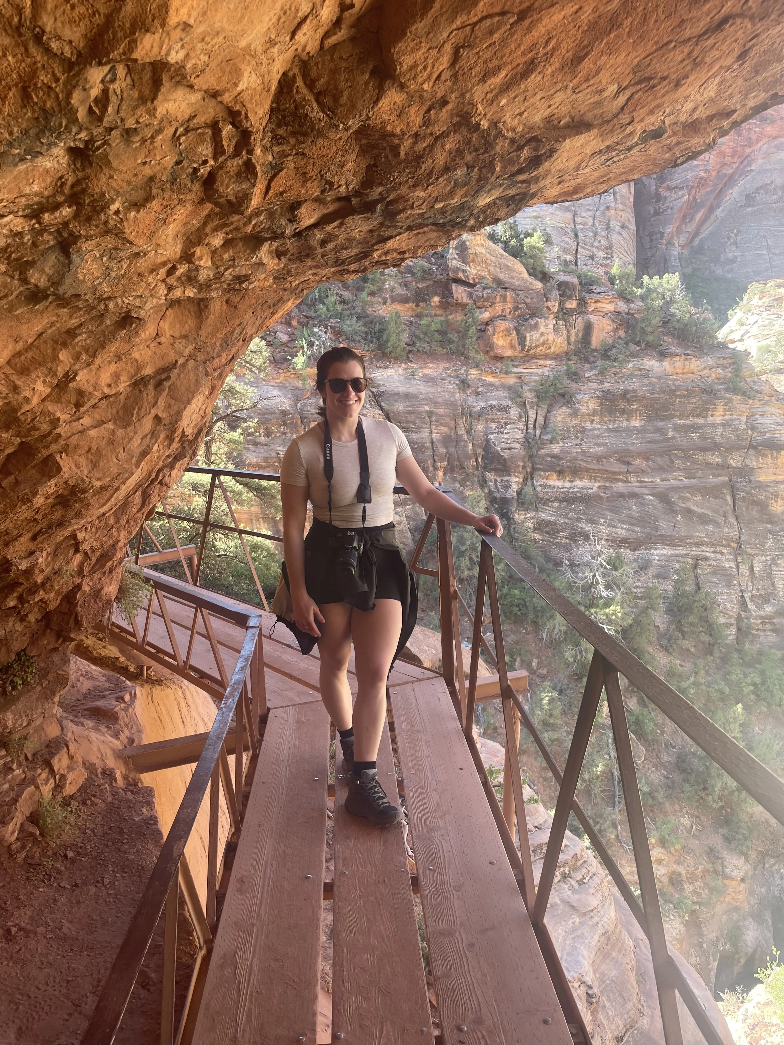 A woman wearing sunglasses, a beige shirt, and black shorts stands on a wooden and metal walkway inside a red rock canyon with a scenic background of cliffs, trees, and a hazy sky.