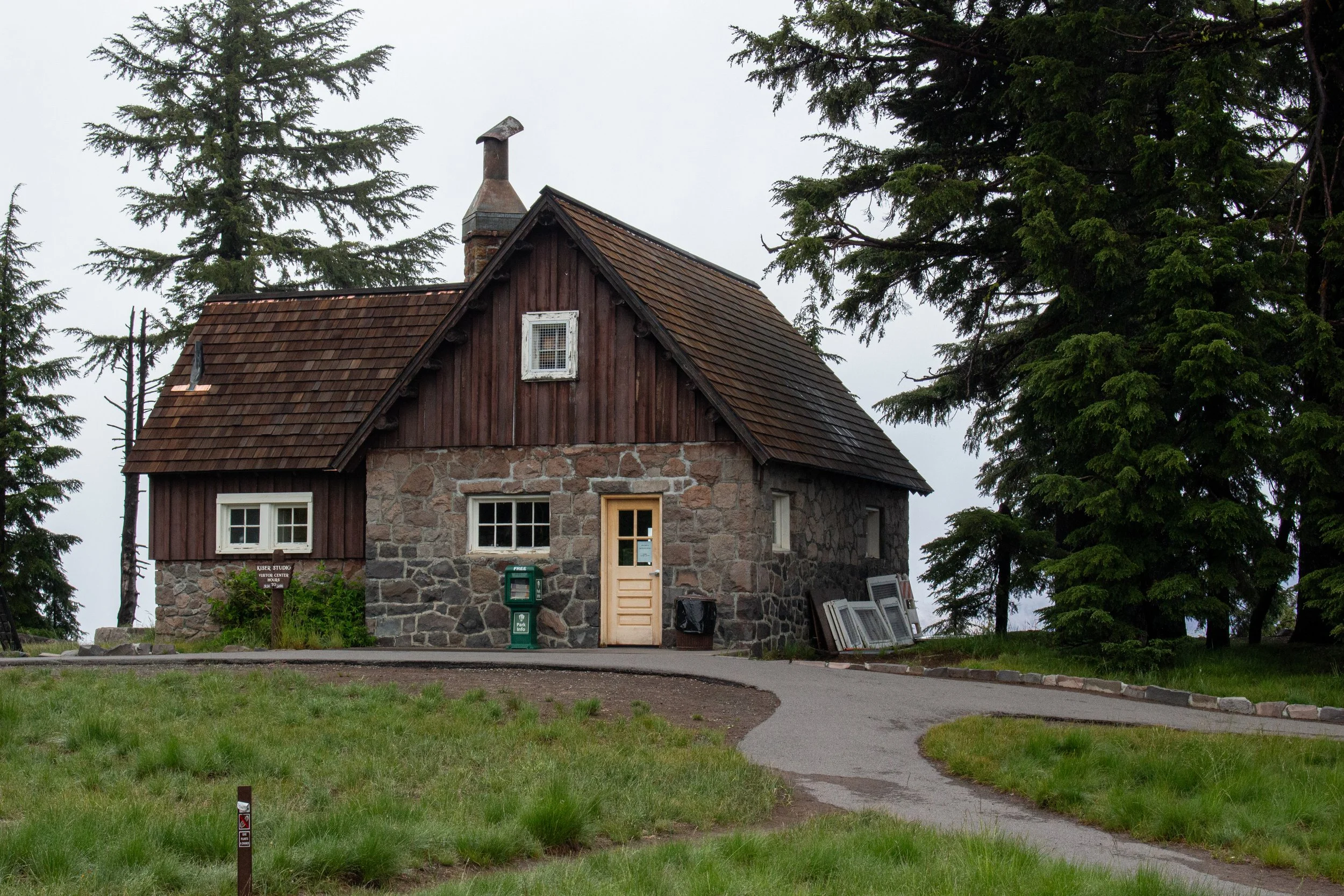 A rustic stone and wood house with a sloped roof during overcast weather, surrounded by large evergreen trees, with a small driveway and grass in the foreground.