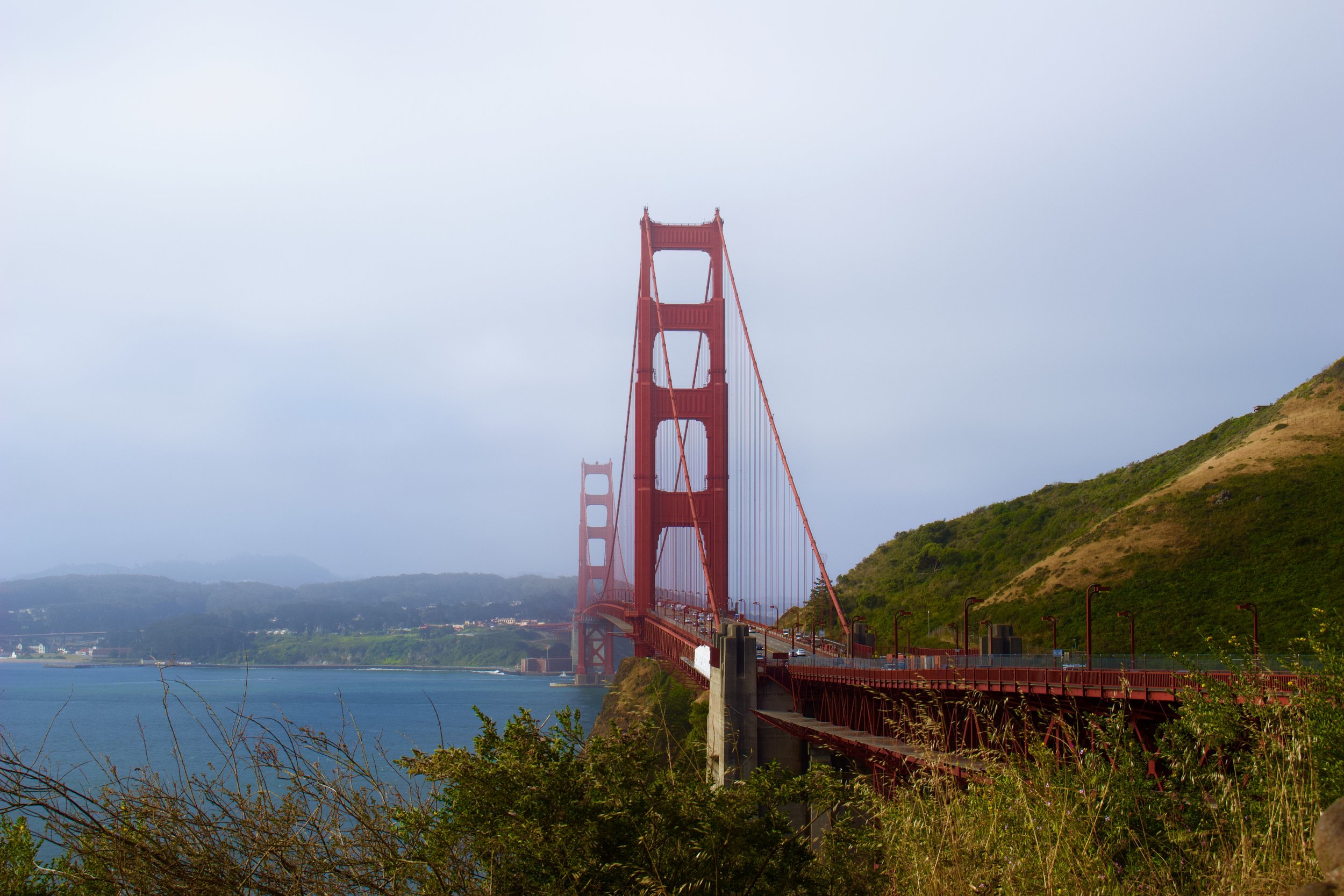 The Golden Gate Bridge, painted in red, spans across the water with foggy sky above and green hills on the right side.