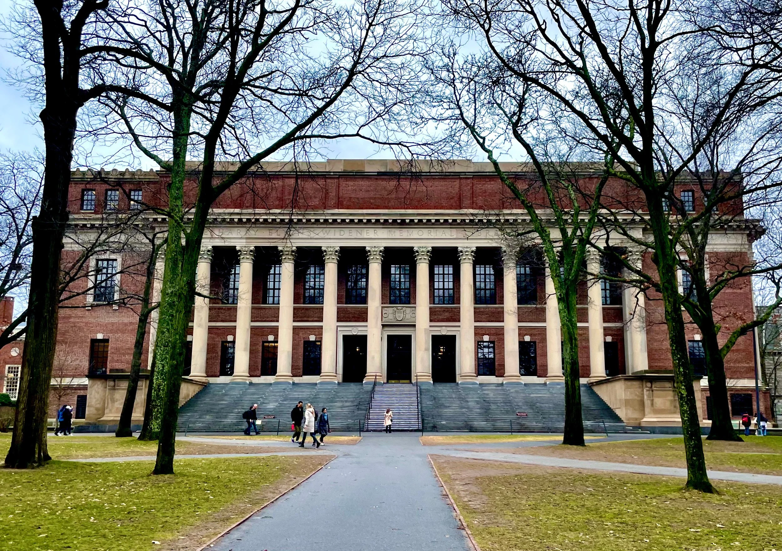A large red brick building with white columns and a portico, surrounded by leafless trees, with a staircase leading up to the entrance and people walking at the front.