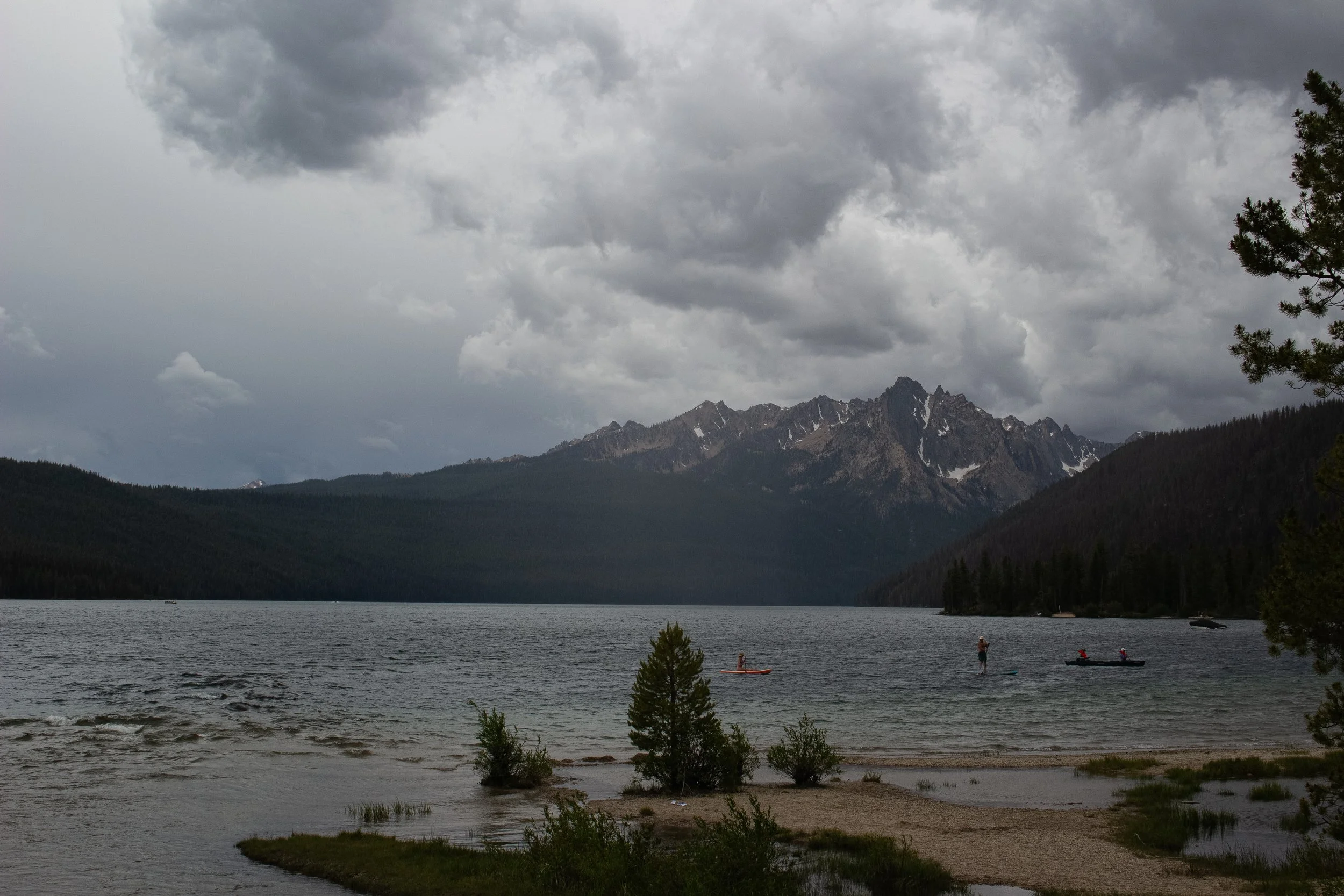 A scenic mountain lake with dark clouds in the sky, surrounded by pine trees, with people canoeing on the water.