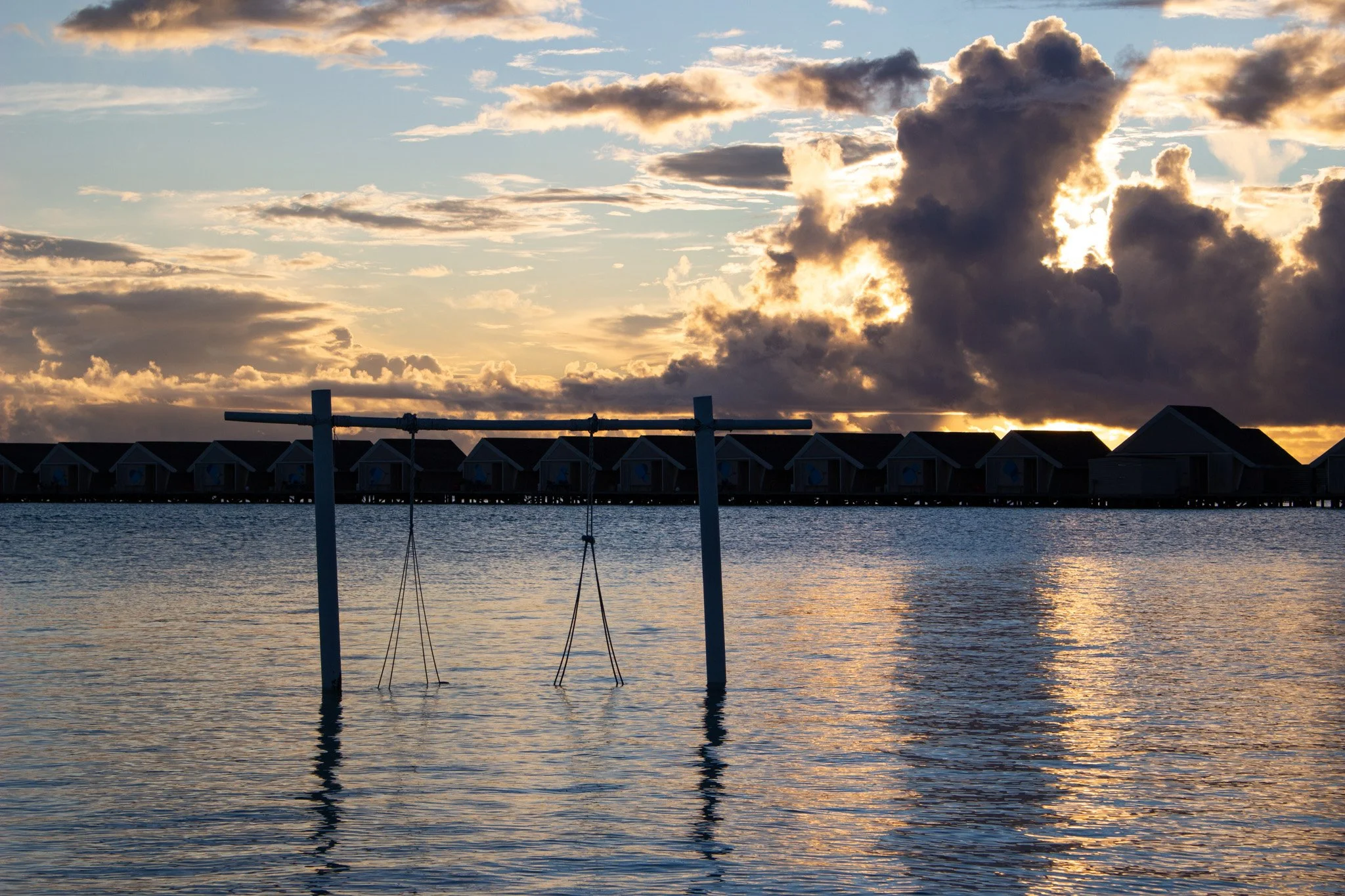 Sunset over water with a row of overwater bungalows in the background and a partially visible building in the foreground.