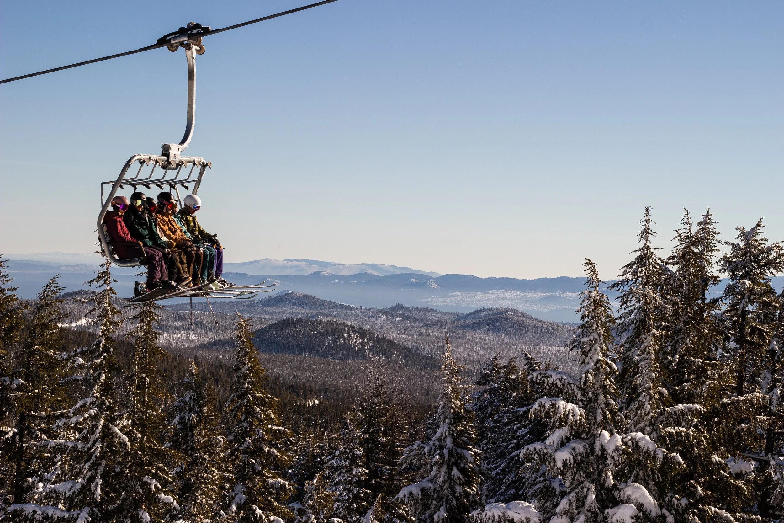 A group of people riding a ski lift in a snowy mountain landscape with pine trees and distant mountains under a clear blue sky.