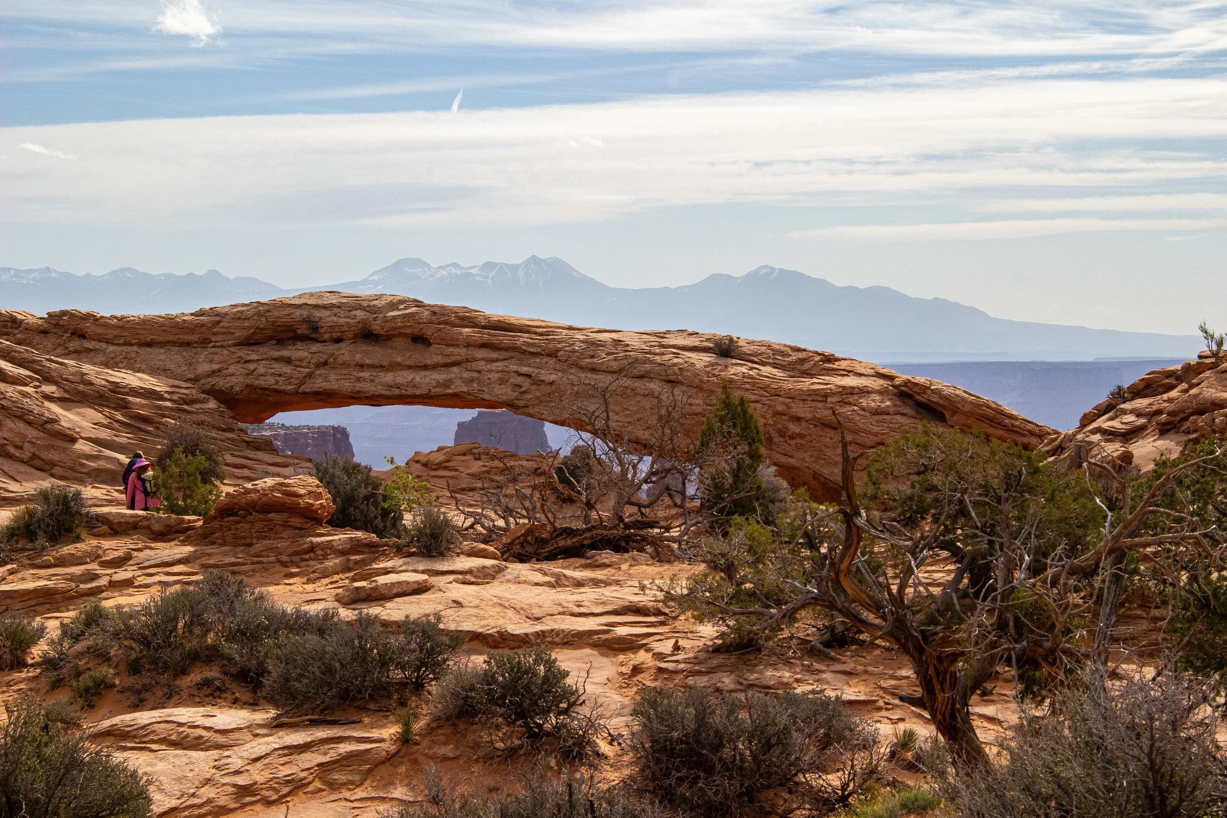 Desert landscape with a natural rock arch, sparse vegetation, and mountains in the background under a partly cloudy sky.