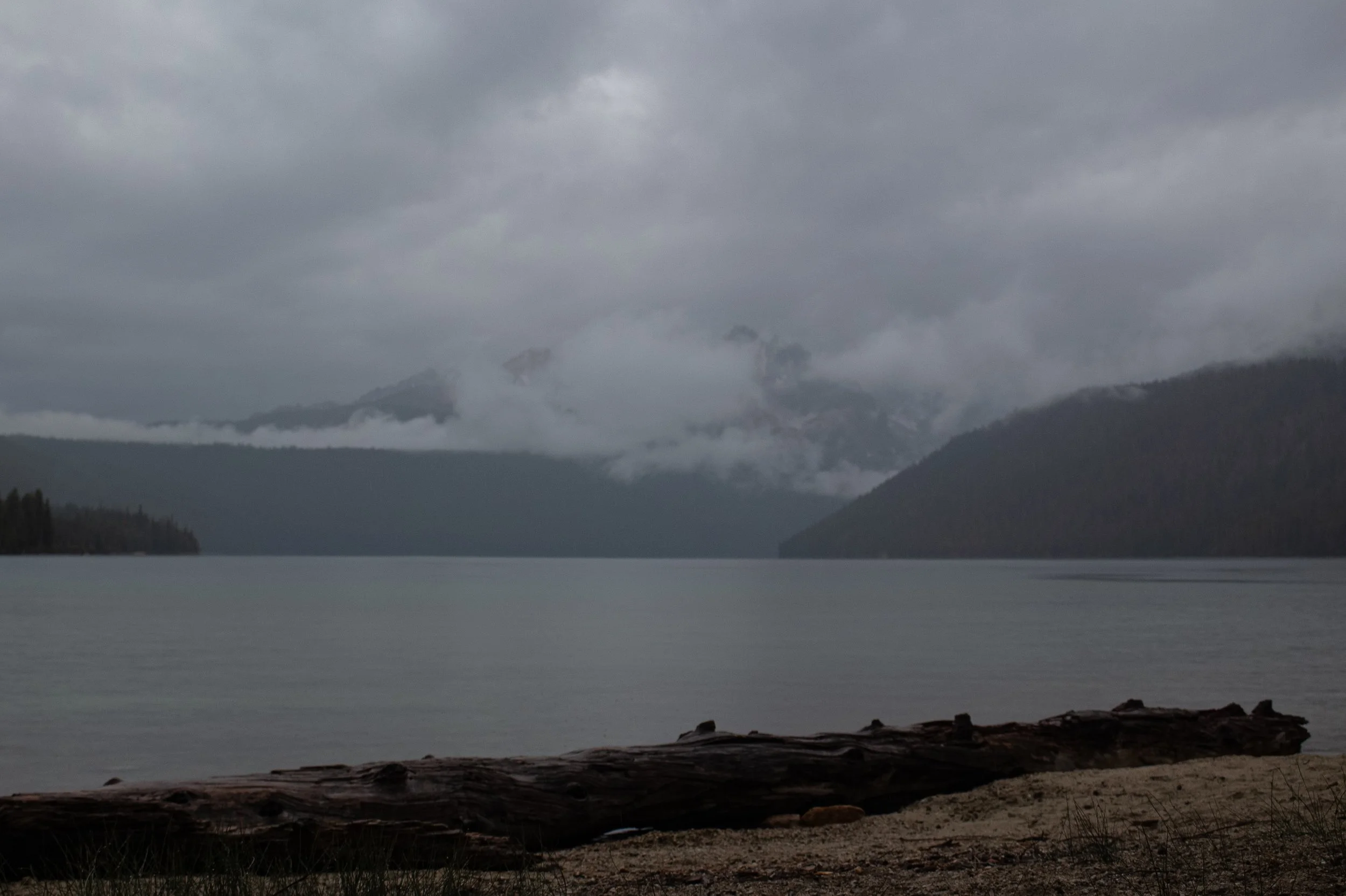 A cloudy, overcast scene of a large lake or river with a mountainous background partially obscured by low-hanging clouds and mist. The shoreline in the foreground features dark rocks and some grass.