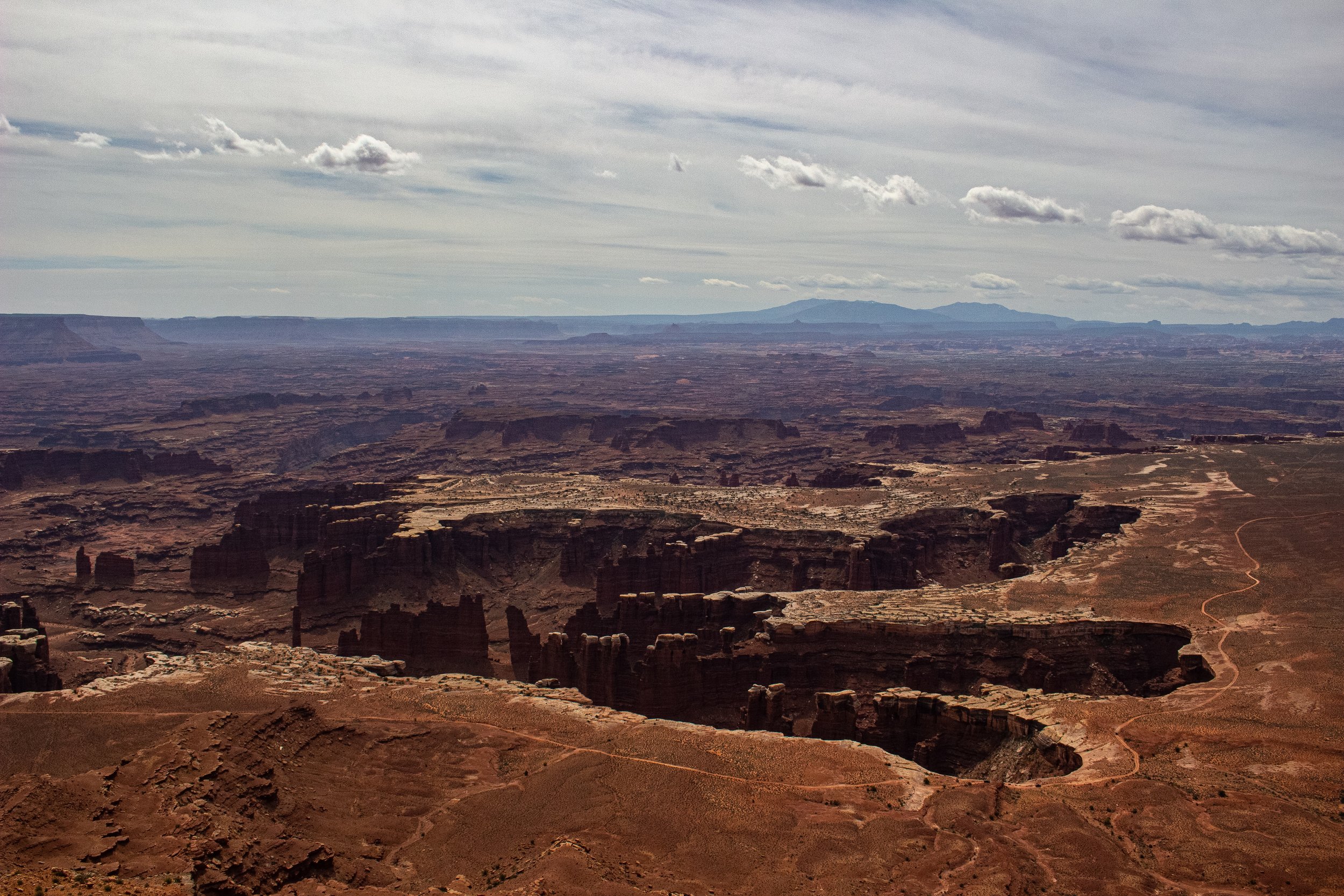 A vast desert landscape with layered rock formations and cliffs, under a cloudy sky with mountains in the distance.