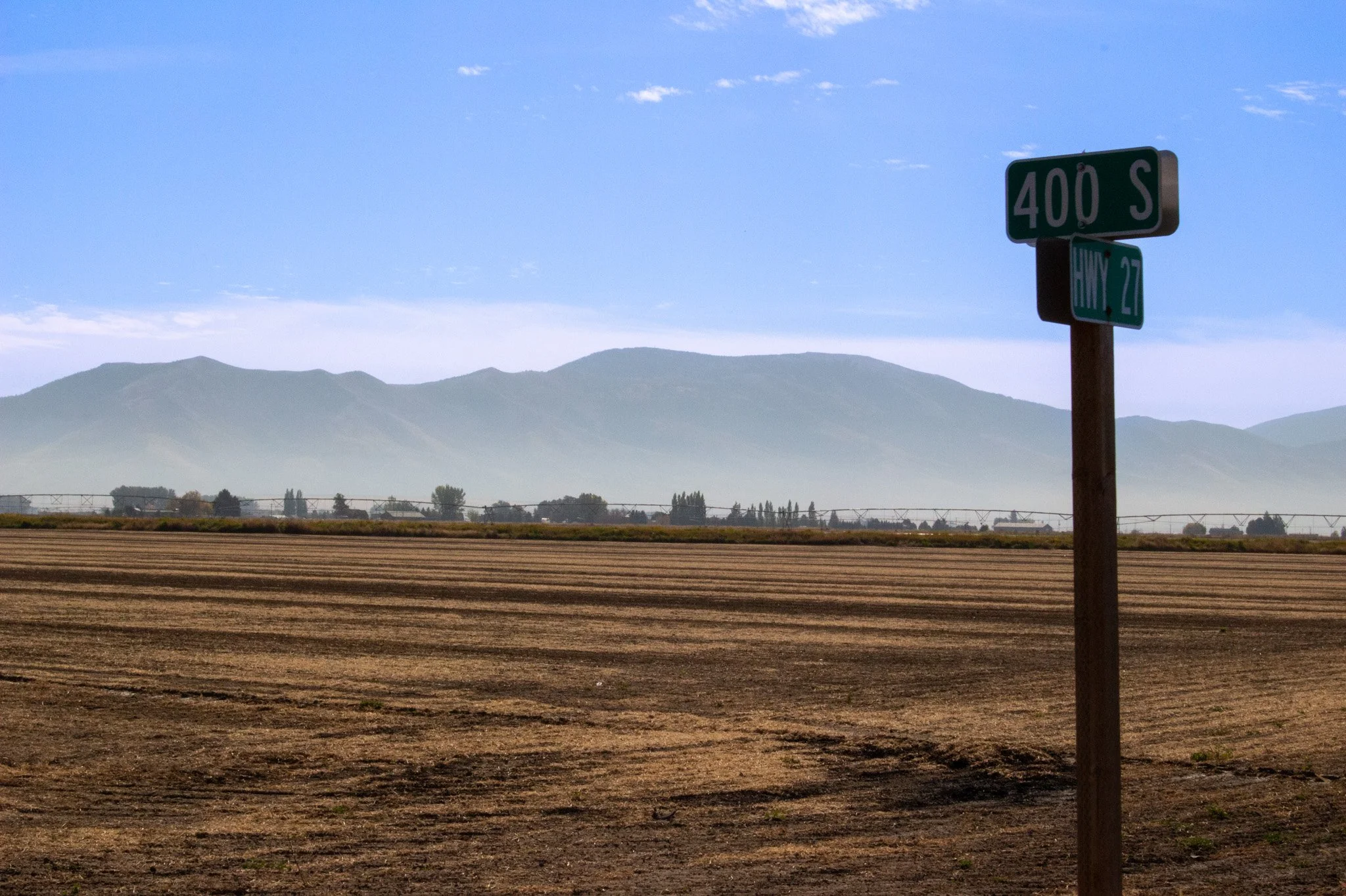 A rural landscape with a dirt field, distant mountains, trees, and a sky with a few clouds. There are two street signs, one indicating 400 S and the other indicating Hily 27.