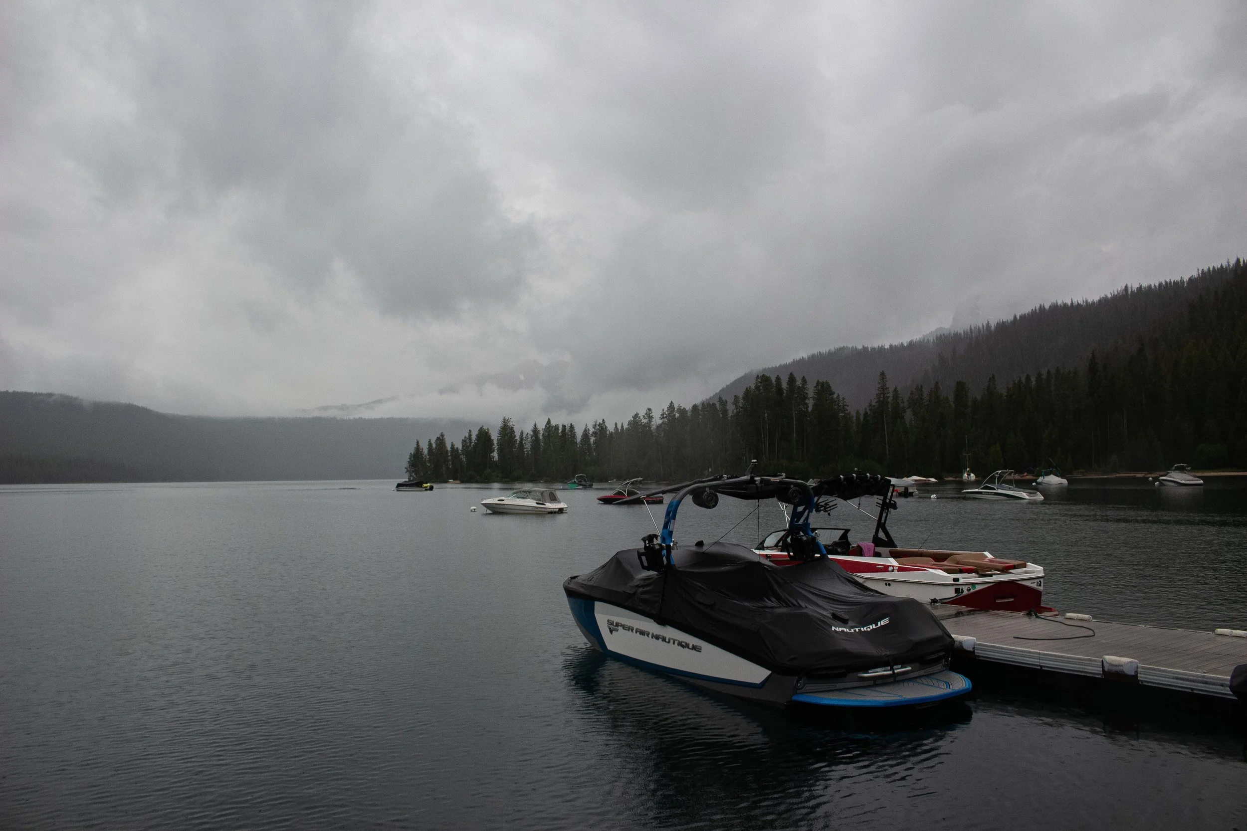 Overcast day at a lake with multiple boats docked along the shore, surrounded by forested mountains.