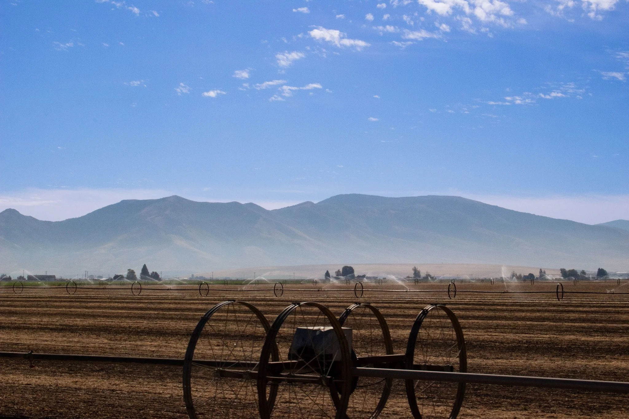 Landscape of farmland with soil fields and irrigation sprinklers, distant mountains under a partly cloudy blue sky.