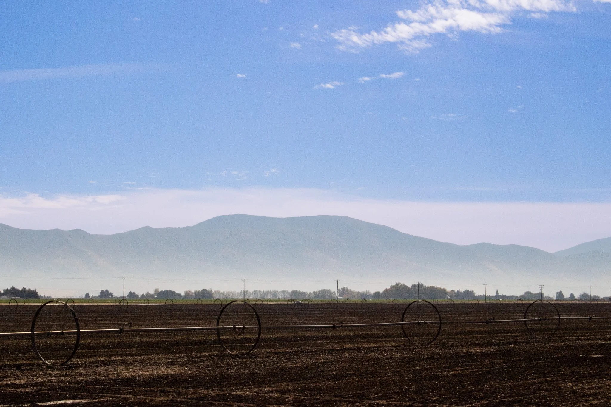 Farmland with circular irrigation systems, mountains in the background, and a blue sky with scattered clouds.