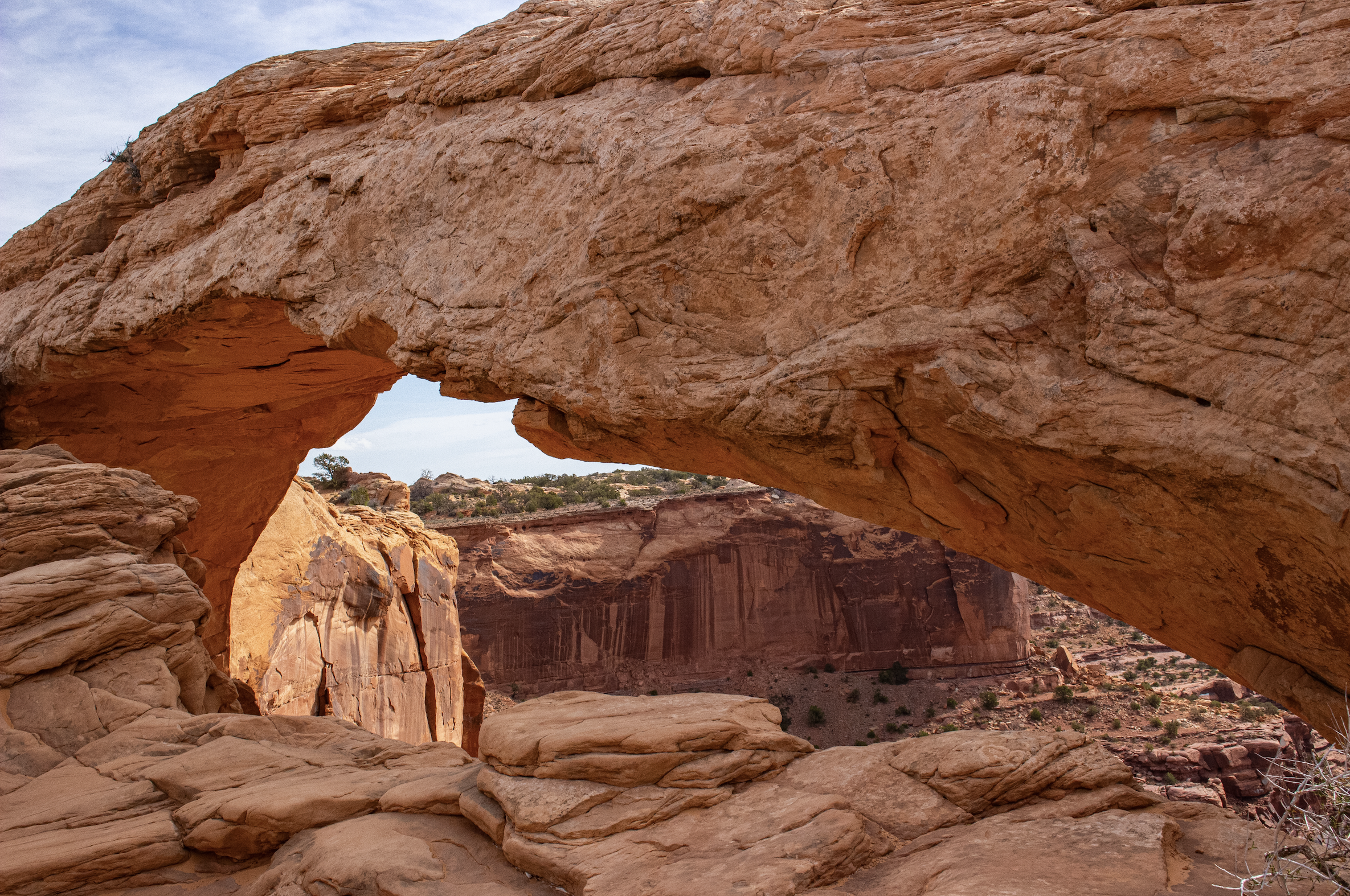 Red rock formation with natural arch in a desert landscape, with sparse vegetation and a cloudy sky.