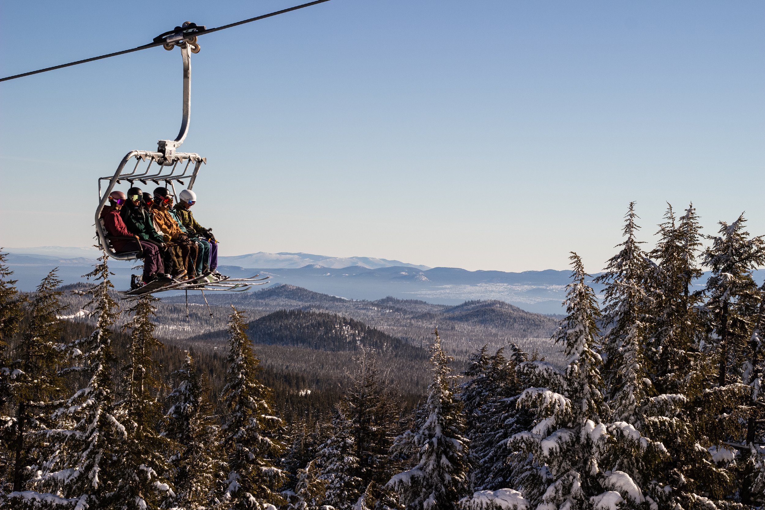 A group of people wearing helmets and winter clothing sitting on a ski lift above a snow-covered forest and mountain landscape.