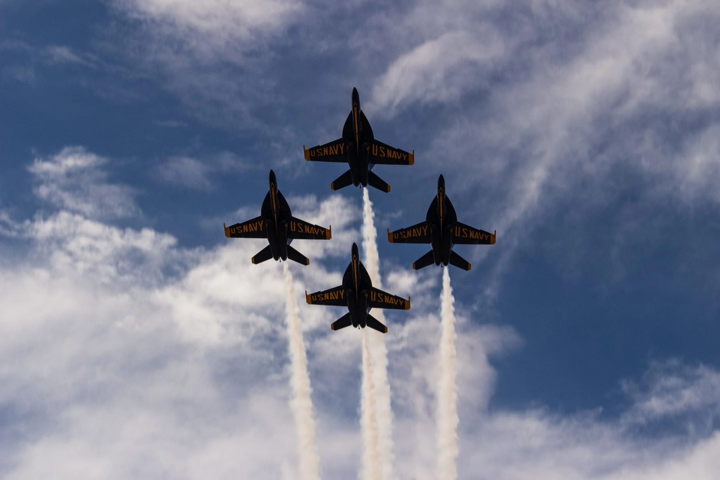 Six U.S. Navy fighter jets flying in a tight V formation in the sky, leaving white smoke trails behind them under a partly cloudy sky.